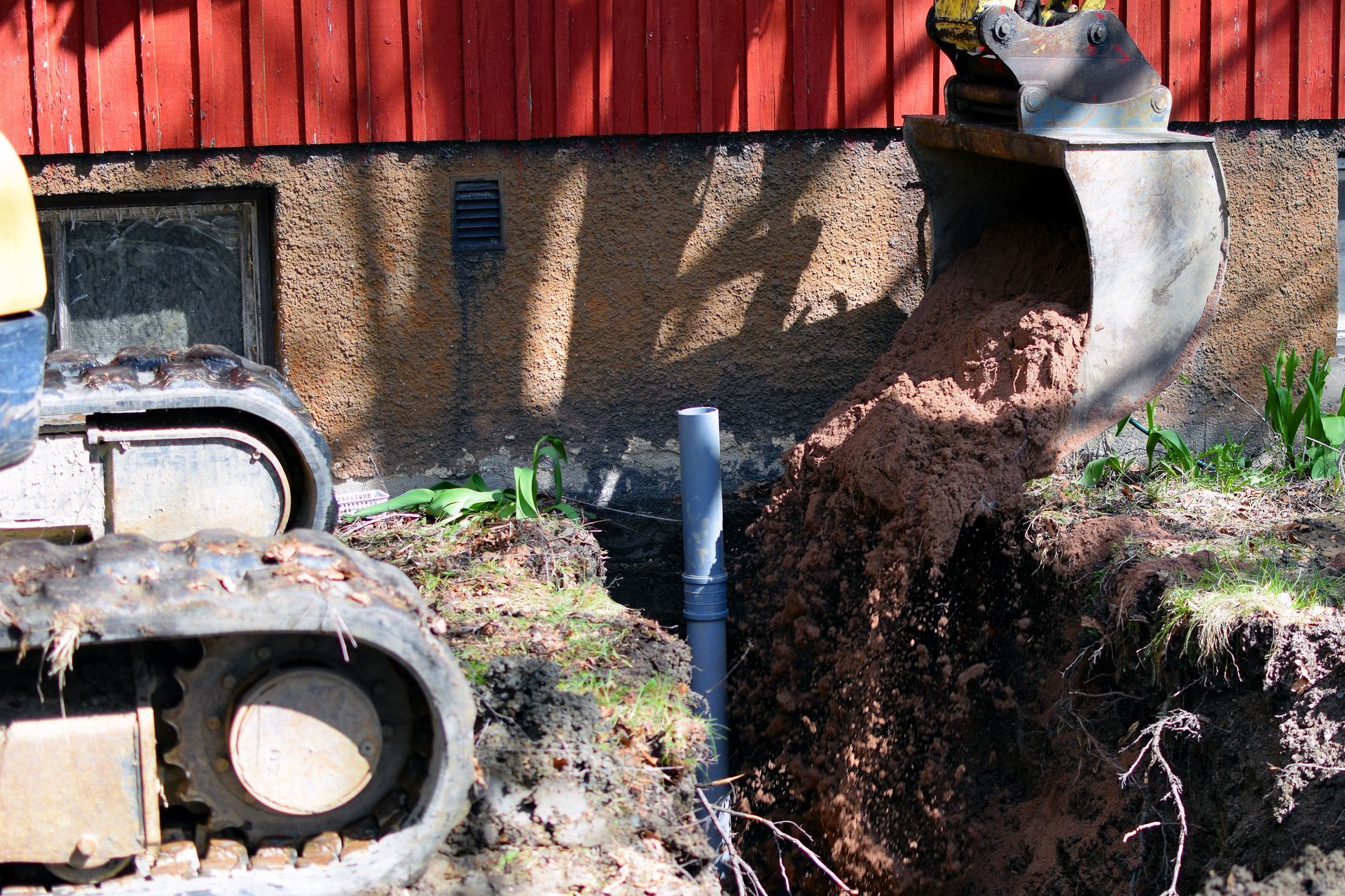 Backhoe digging a trench beside a red building, with dirt piling up on the right