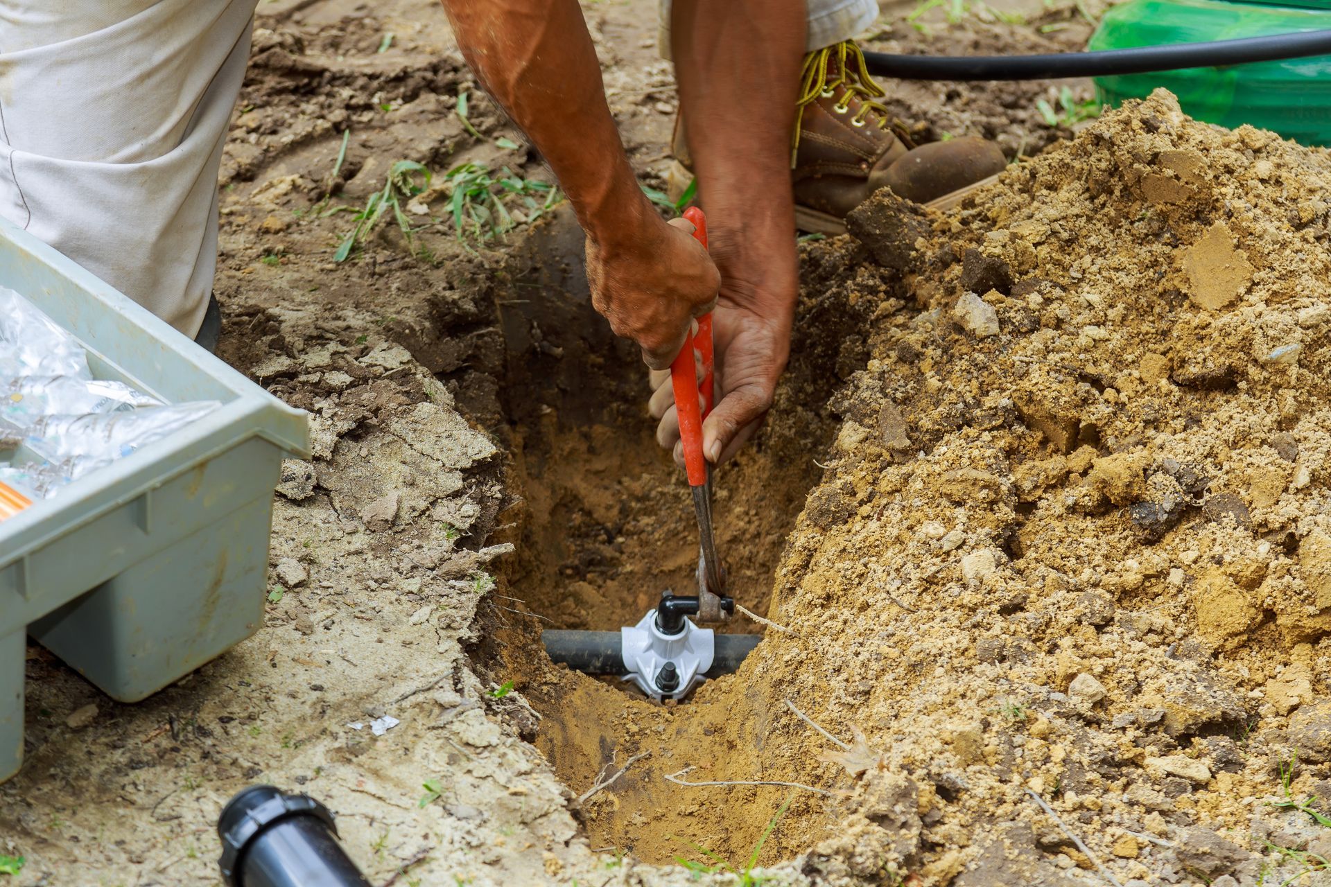 Worker digging in sandy soil with a red-handled tool beside a small trench.