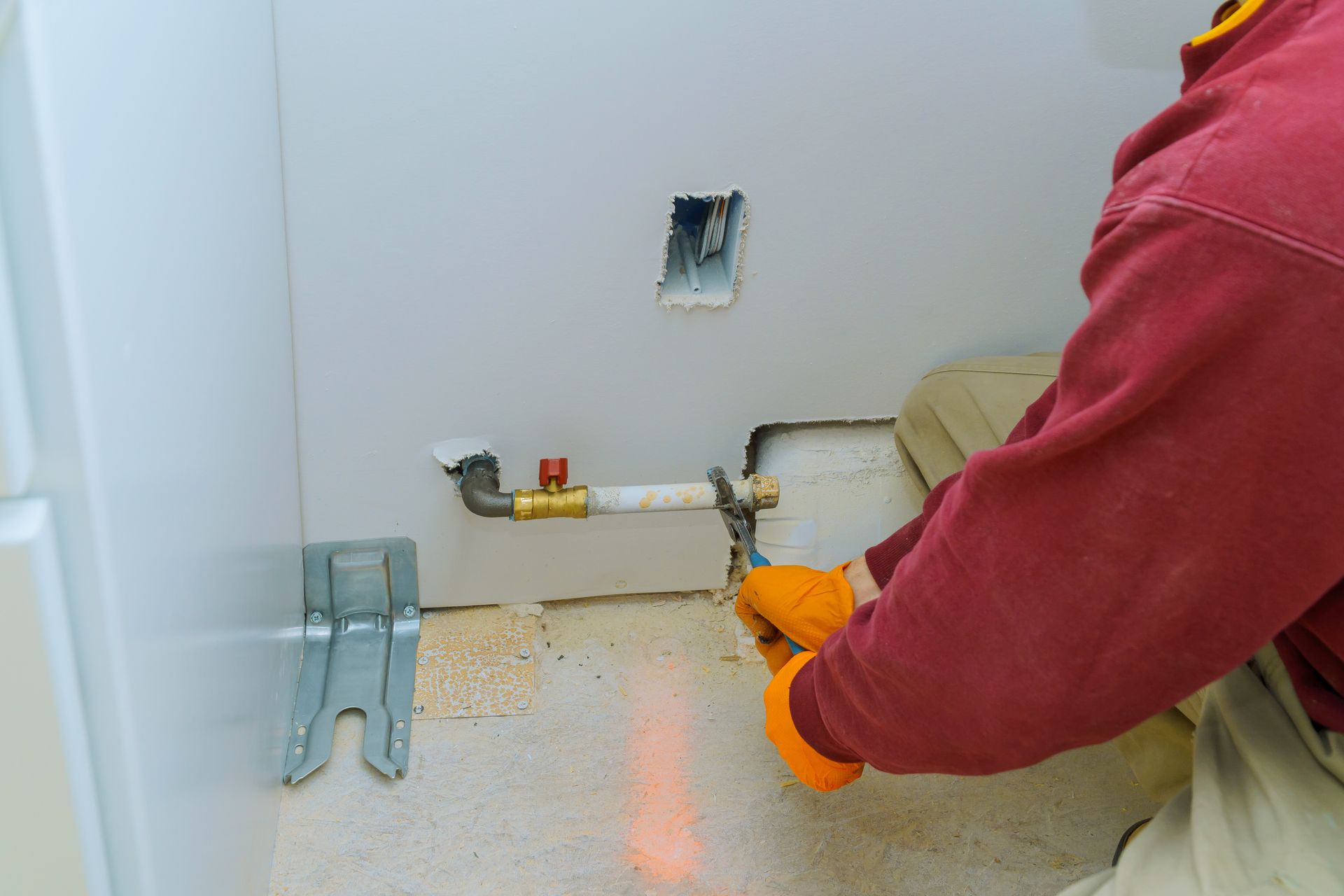 Person in red shirt using orange gloves to work on exposed plumbing near a wall outlet.