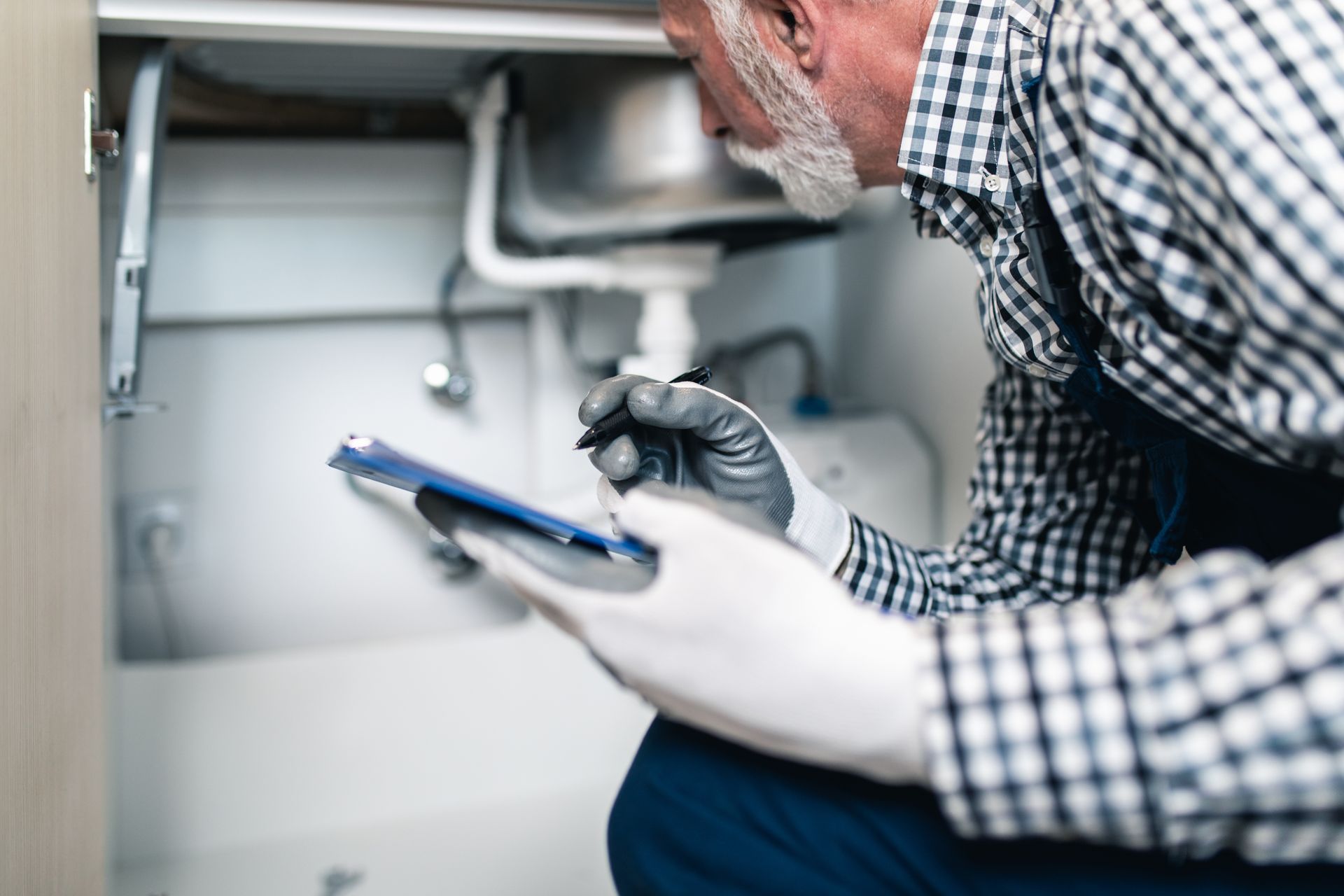 Plumber inspecting under-sink pipes with a flashlight and clipboard in a kitchen cabinet