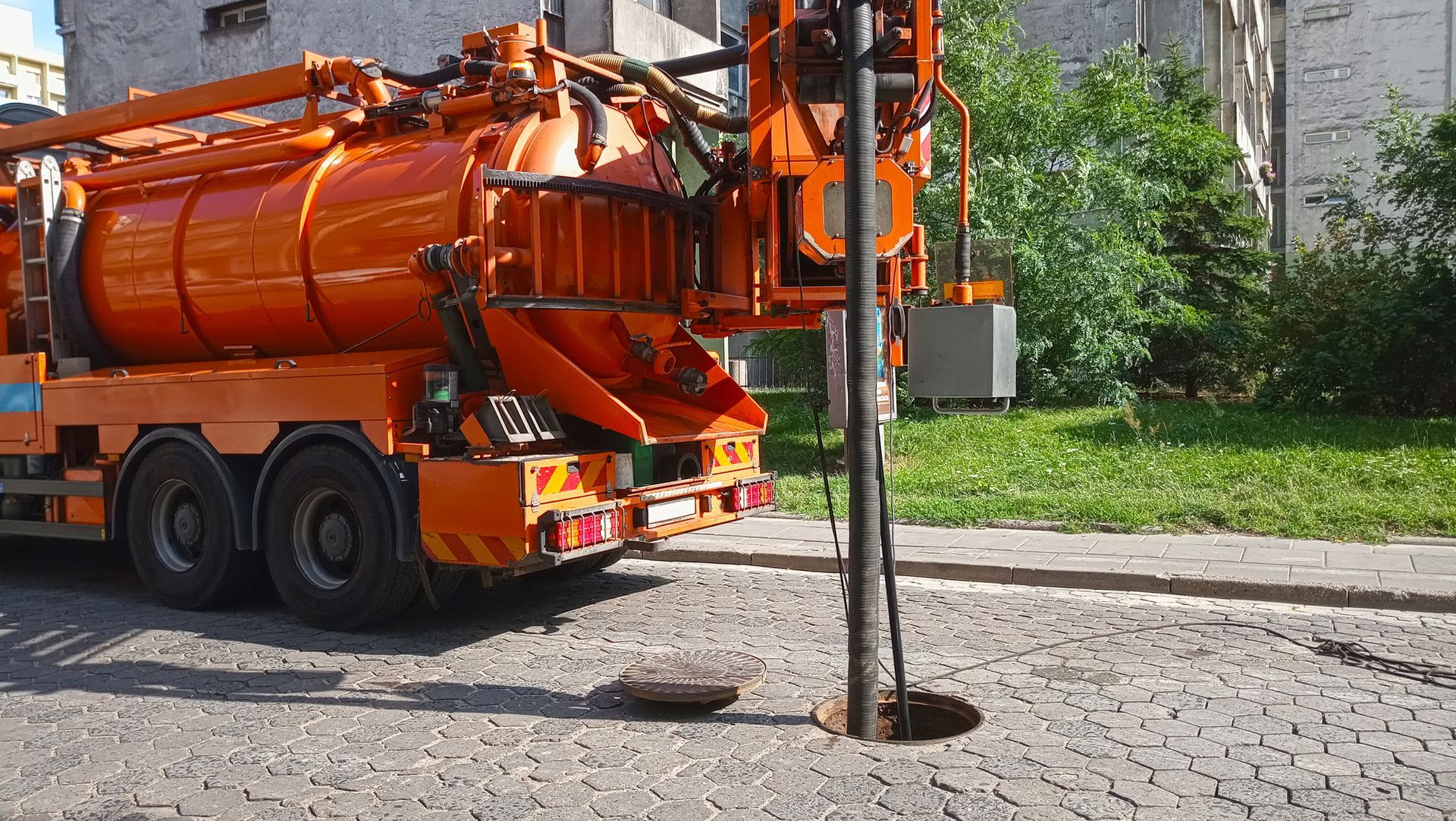 Orange sewer vacuum truck cleaning a street drain with a hose and metal pole on a gravel roadside.