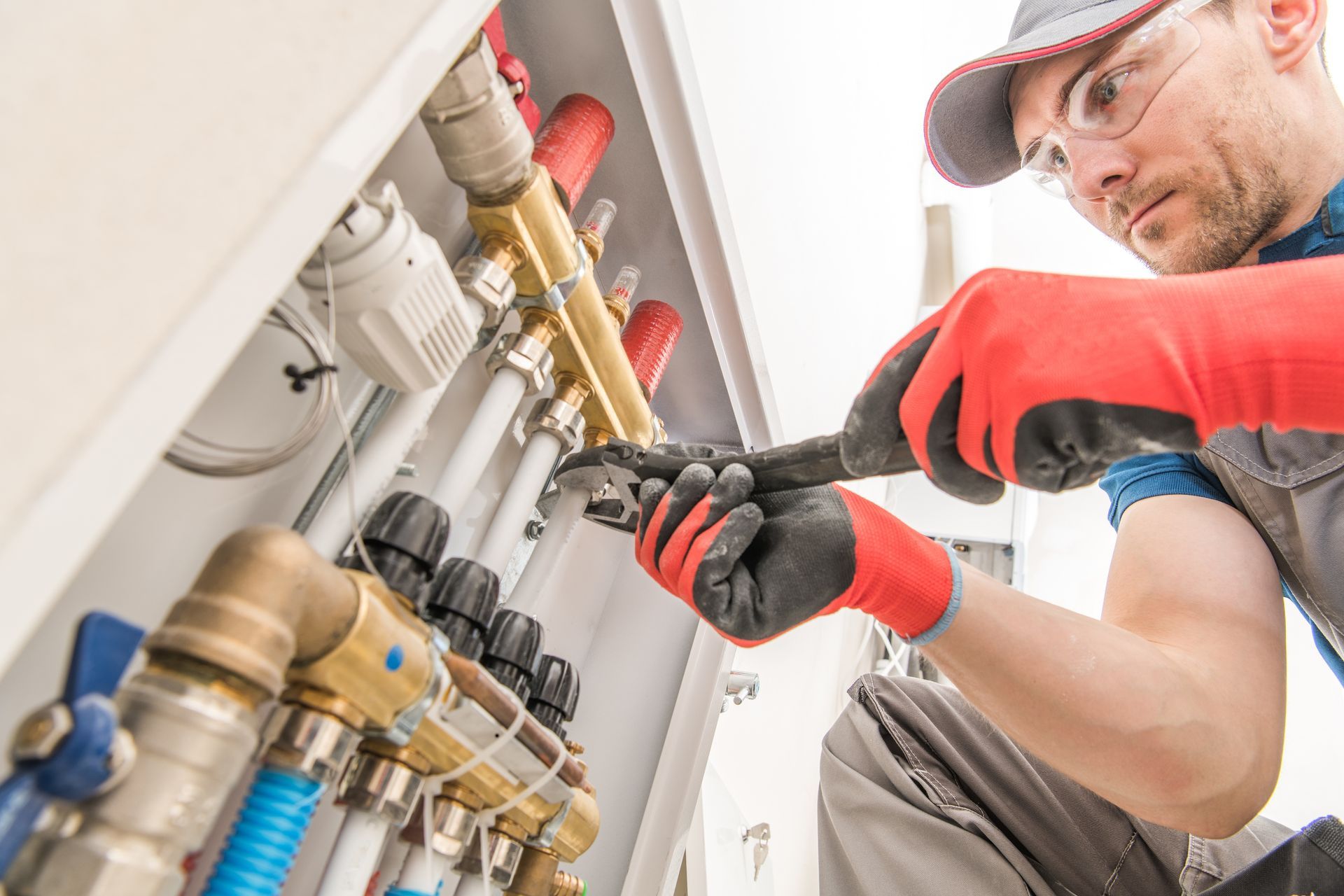 Plumber tightening a pipe connection under a sink with a wrench, wearing red gloves and a cap