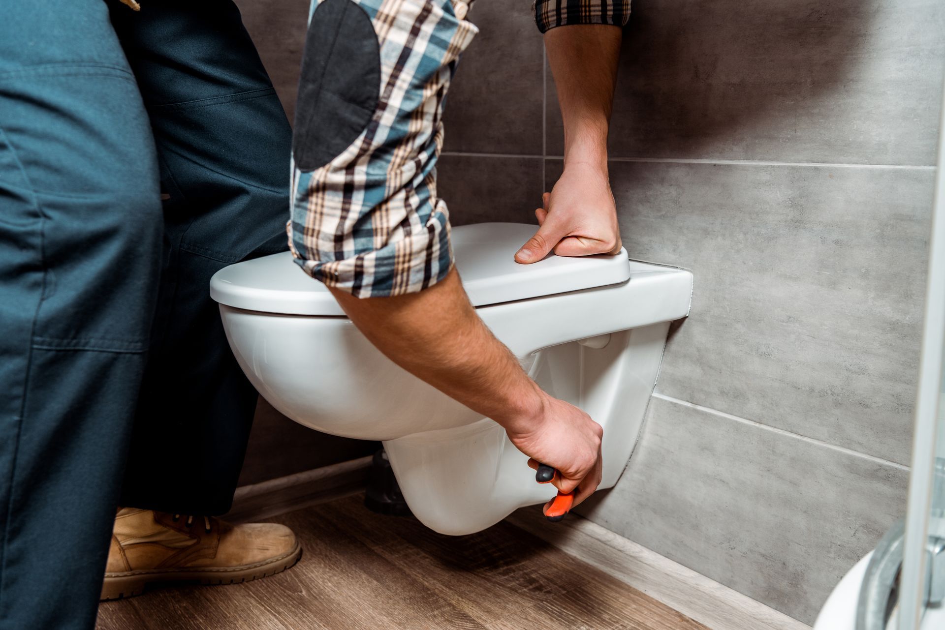 Person installing a white wall-mounted toilet in a tiled bathroom with a wrench
