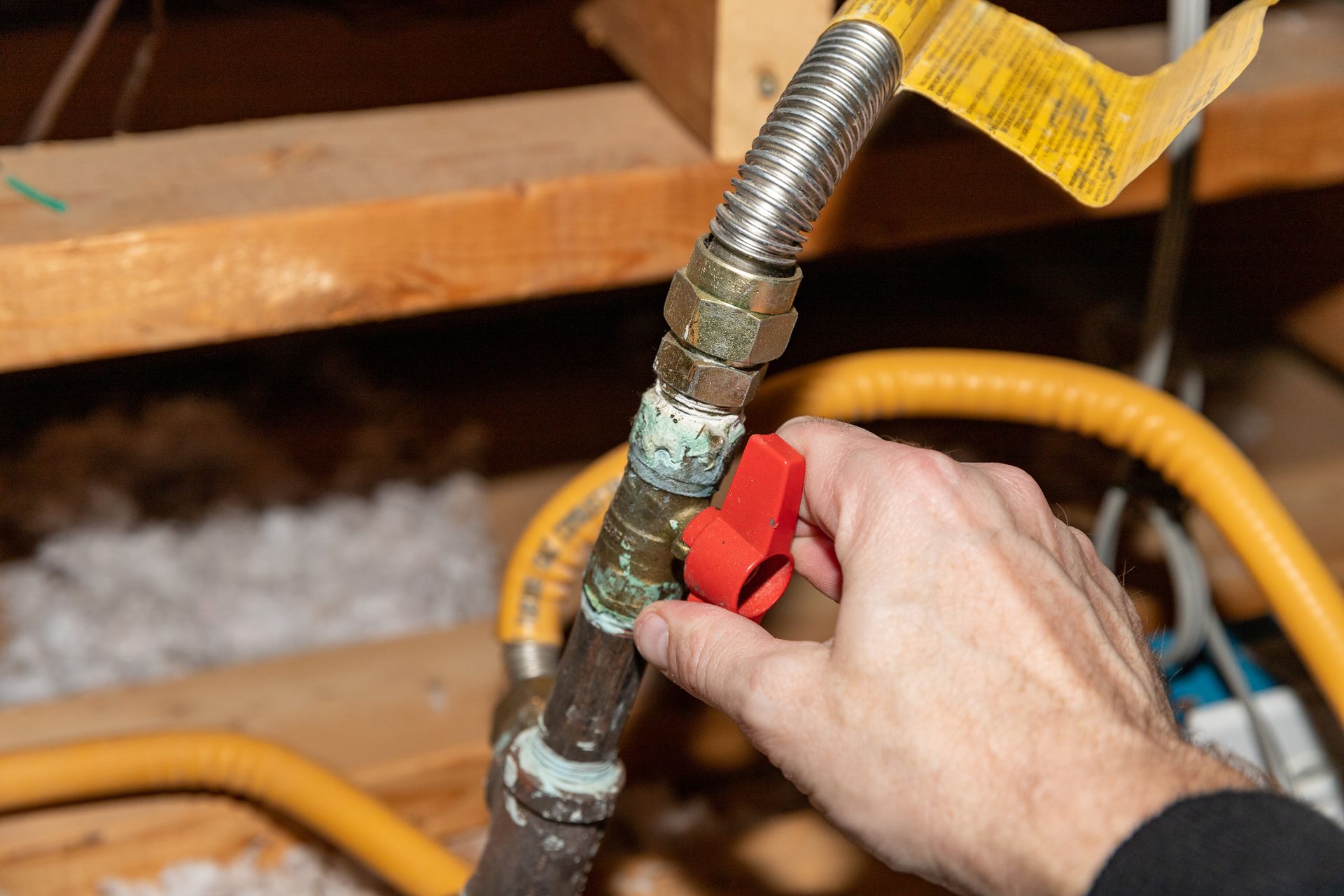 Hand turning a red gas shutoff valve on a pipe with yellow tubing in a basement area
