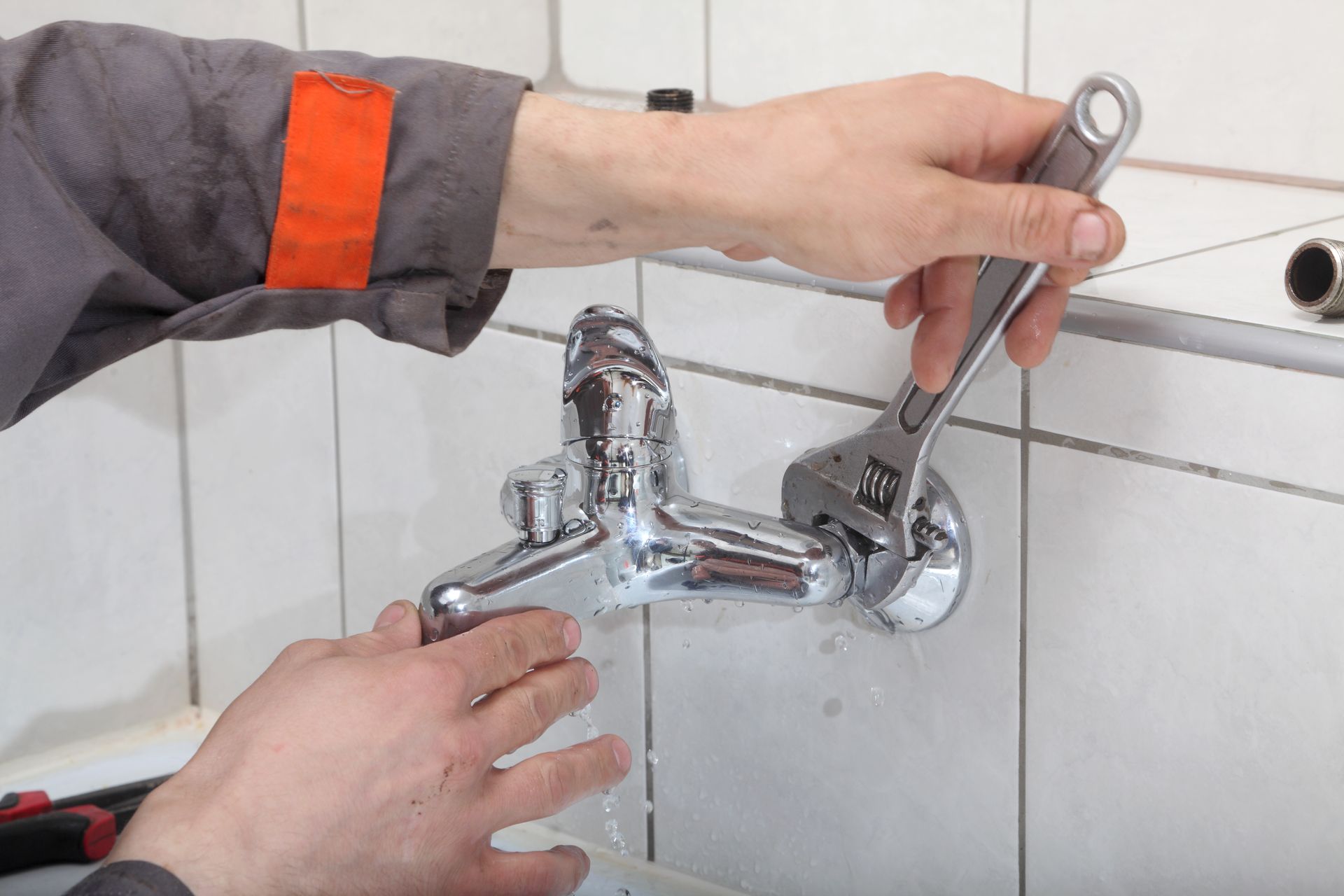 Hands using a wrench to repair a chrome bathroom faucet on white tiled wall