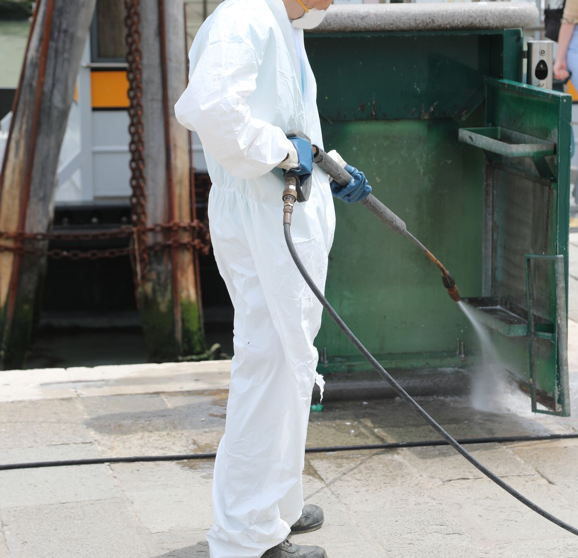 Person in white protective suit pressure-washing a green industrial surface outdoors