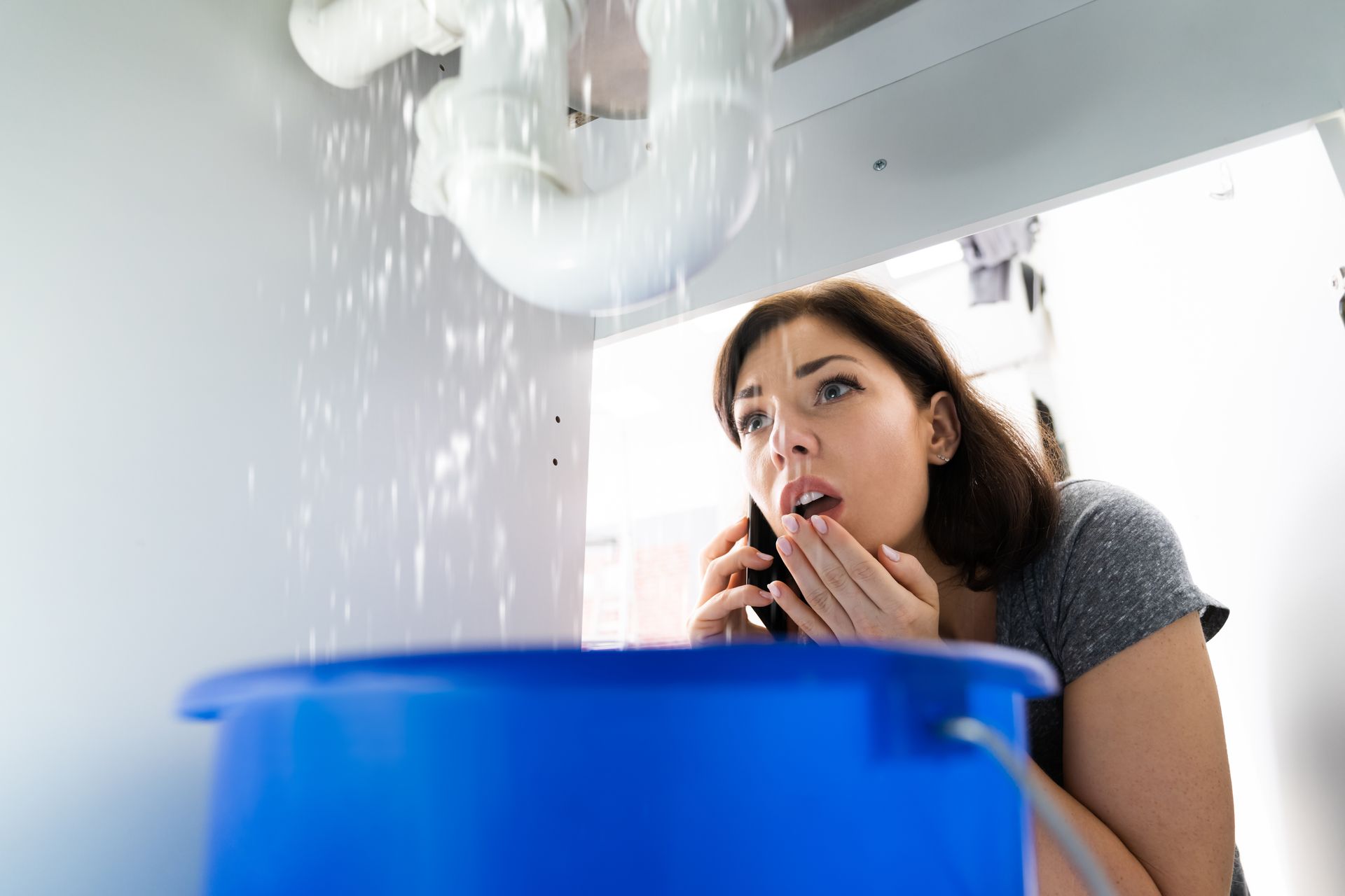 Woman staring at a ceiling leak, holding her hands to her mouth as water drips into a blue bucket