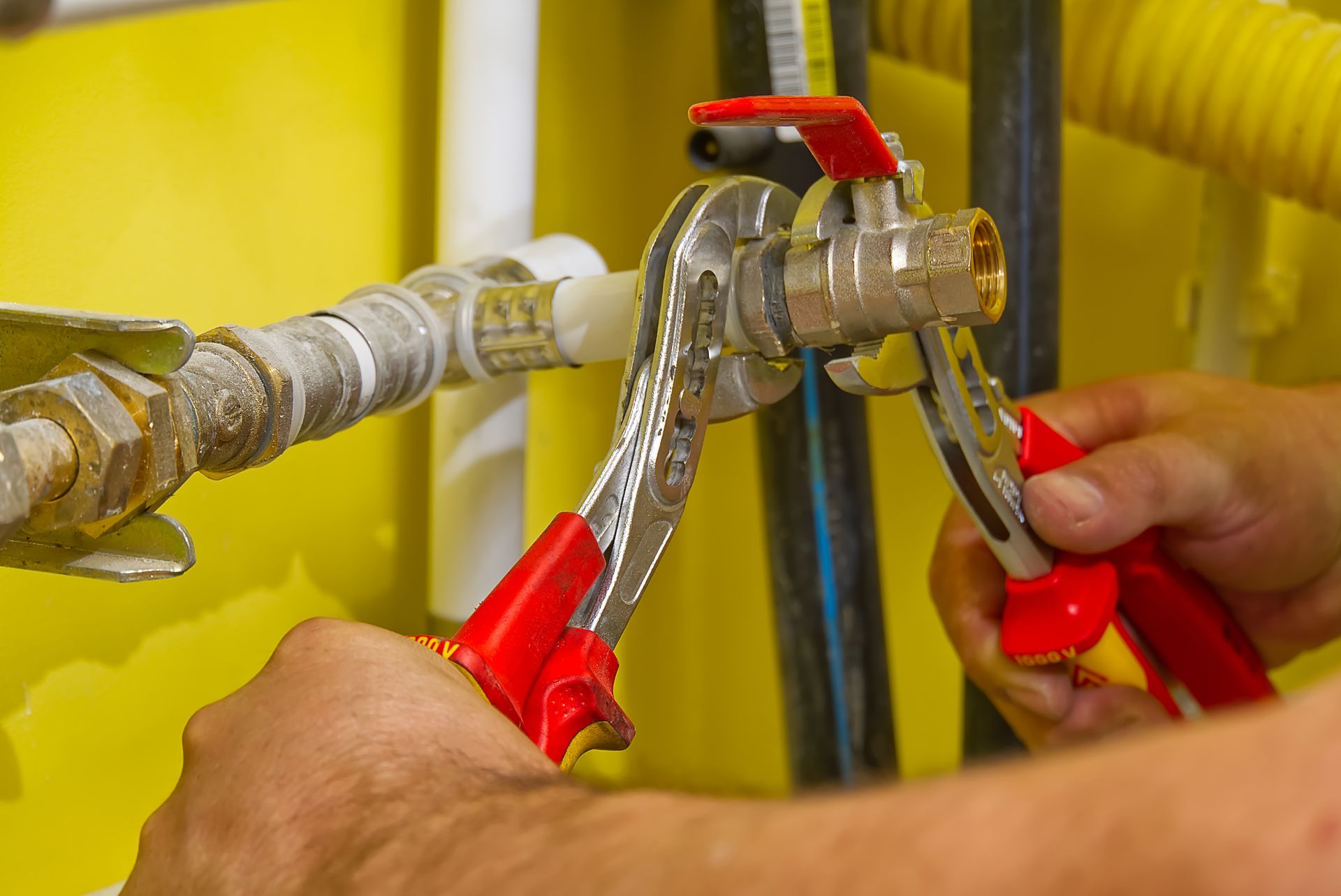 Hands using red-handled pliers to work on a pipe valve with a red shutoff lever, close-up.