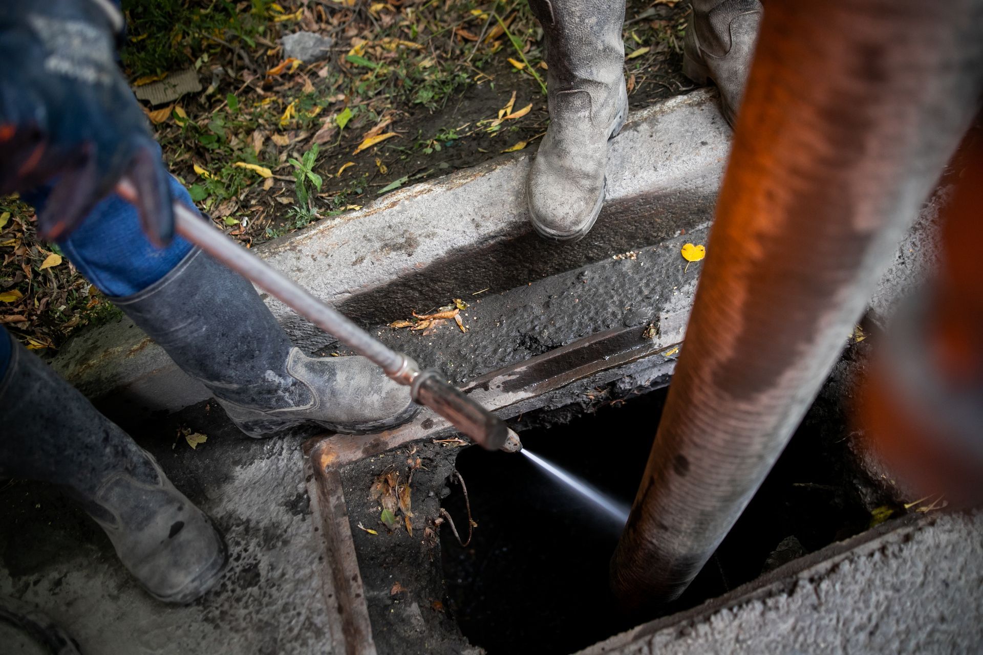 Worker uses a pole to inspect a dark manhole beside a concrete curb and rusty pipe.