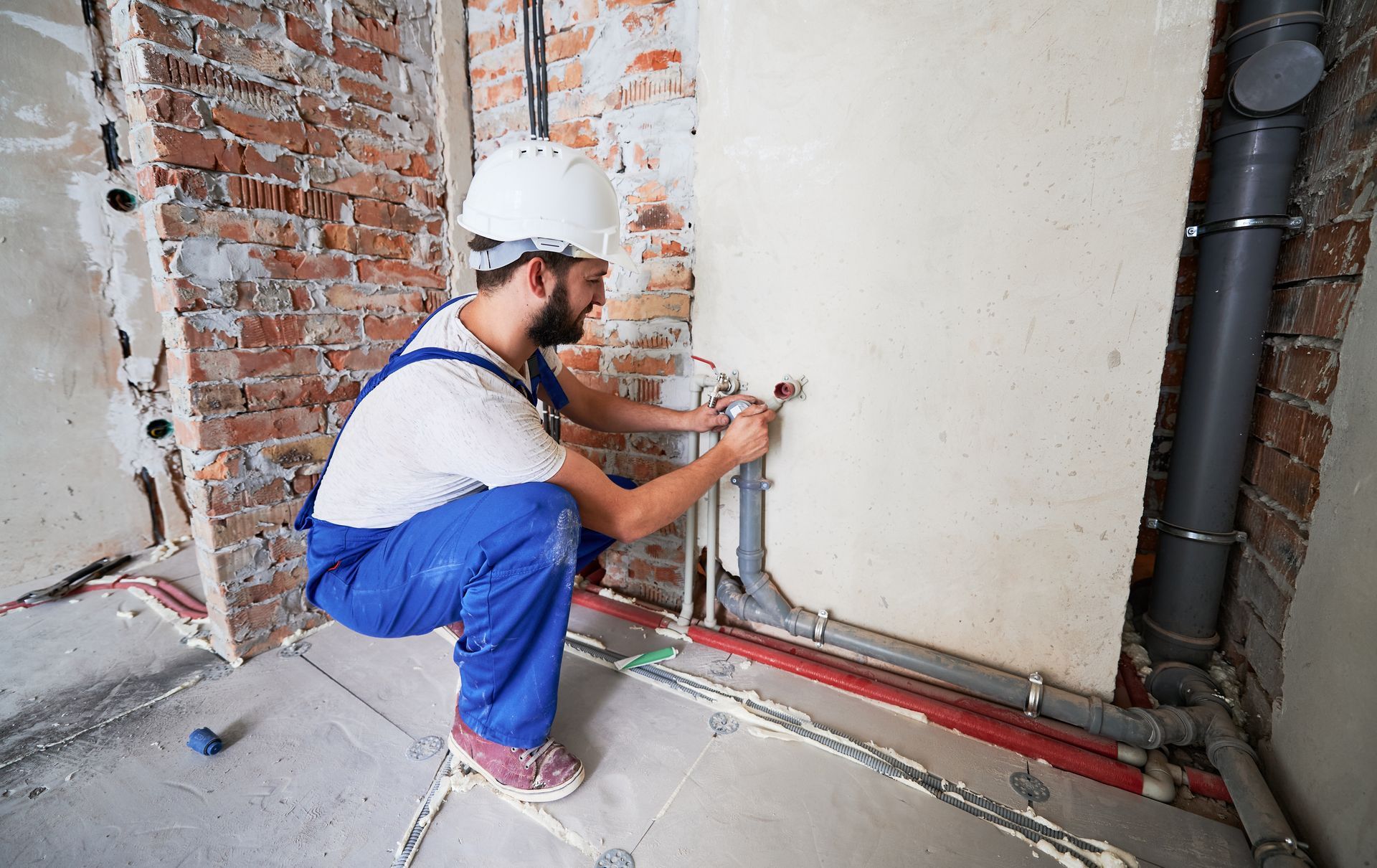 Worker in blue overalls installing pipes on a construction site near a brick wall and exposed plumbing.