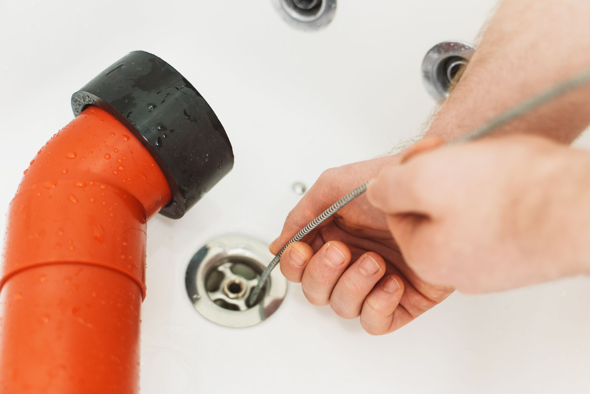 Hand using a drain snake to unclog a white sink beside an orange drainpipe