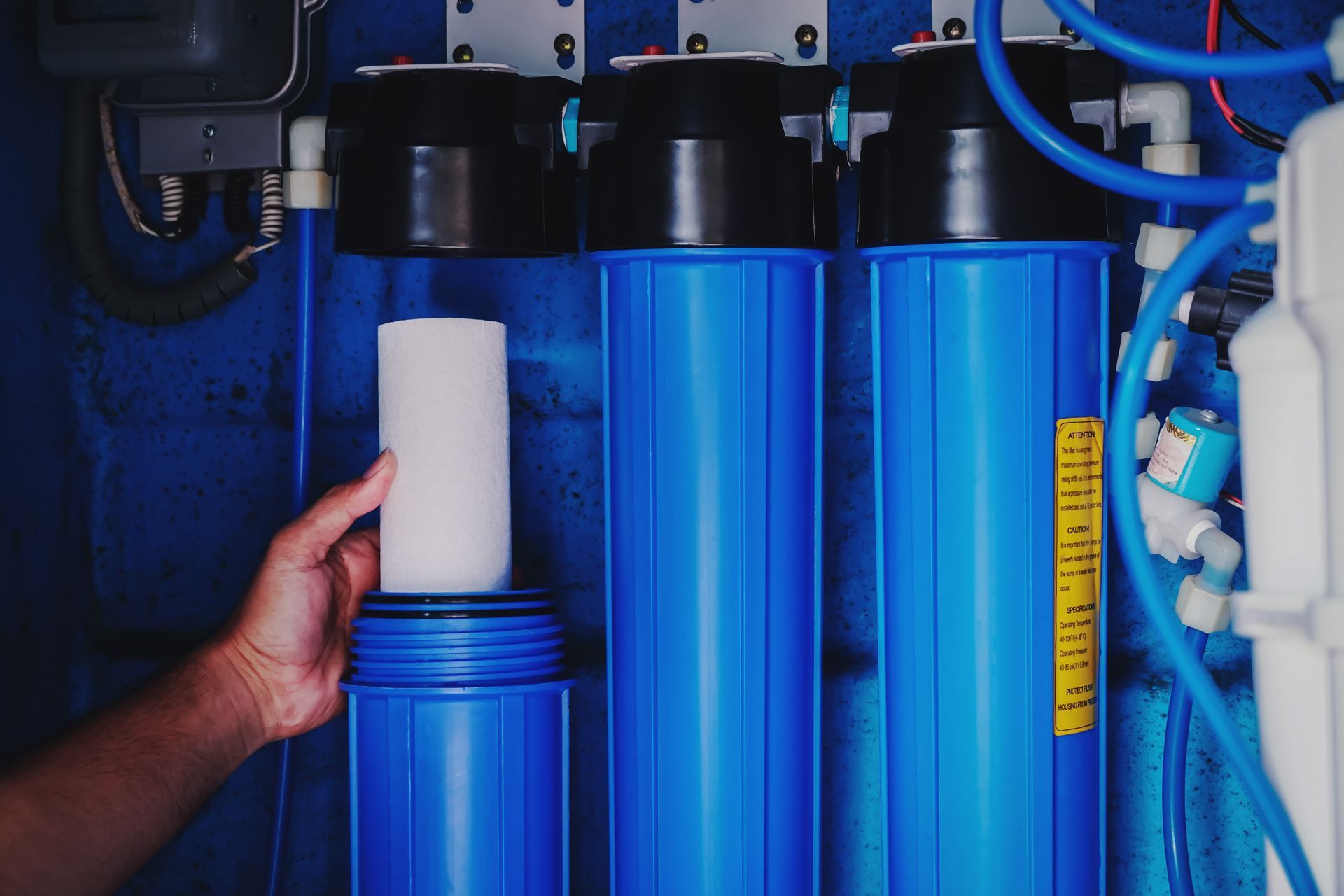 Hand replacing a white filter cartridge in blue water filtration system with pipes and fittings