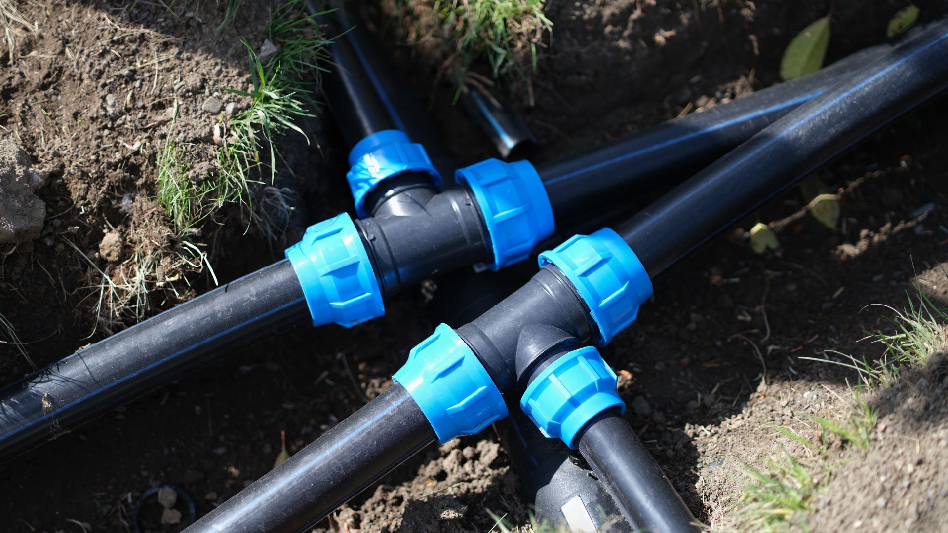 Black irrigation pipes joined by blue plastic fittings in a dirt trench.