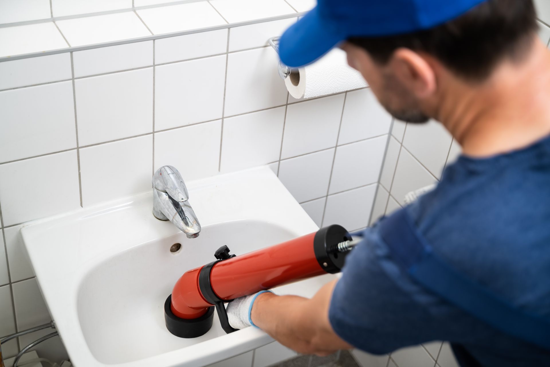 Plumber using a red drain auger to clear a white sink in a tiled bathroom