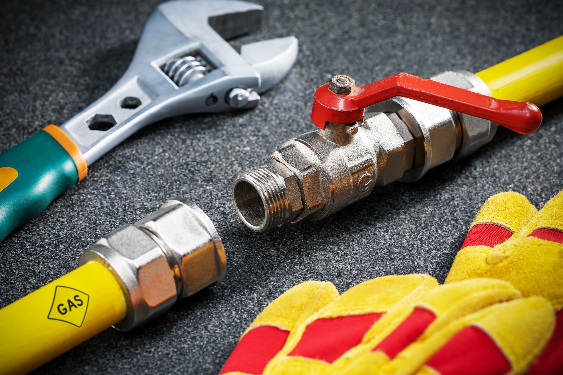 Plumber's tools on a gray surface, including wrenches, pipe fittings, and yellow-red work gloves.