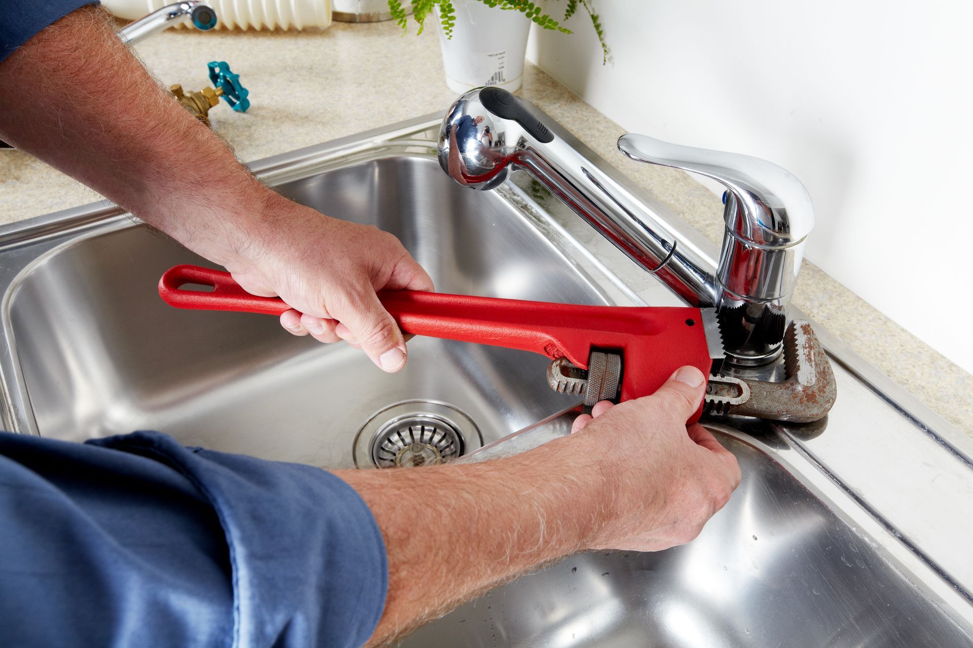Hands using a red pipe wrench to loosen a faucet under a kitchen sink