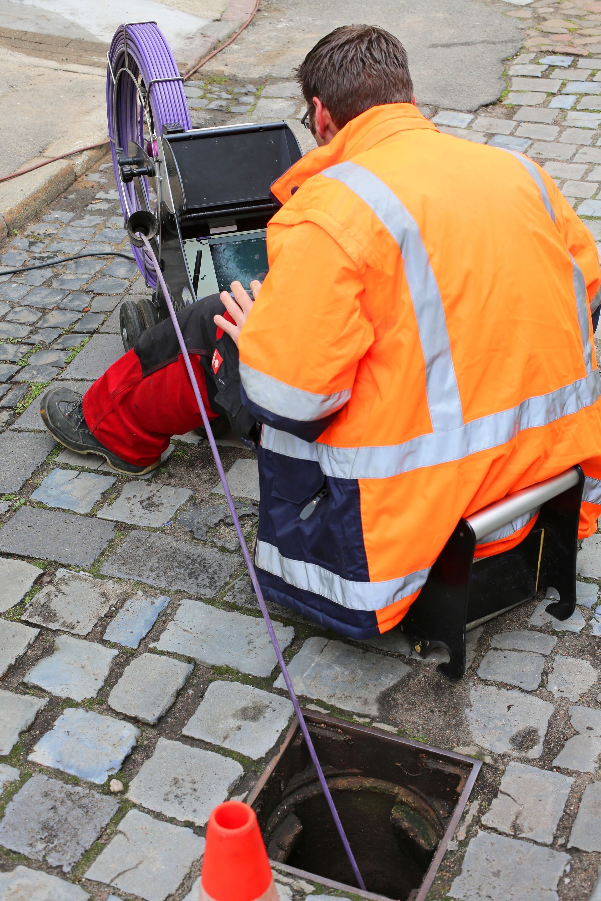 Worker in orange safety jacket inspecting a street drain with equipment near an open manhole.