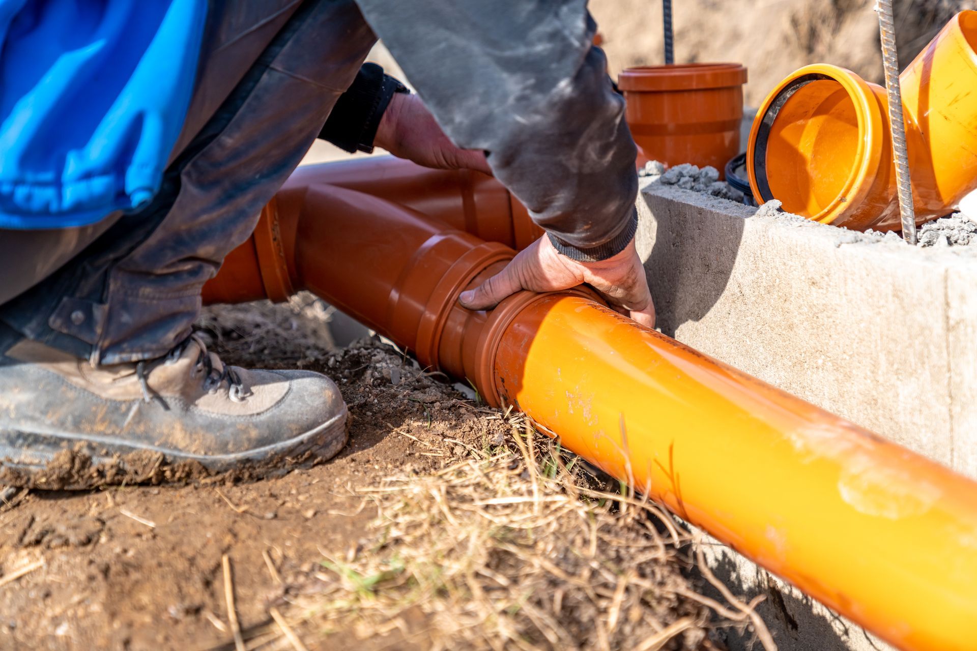Worker fitting orange PVC pipes in a trench next to concrete blocks outdoors