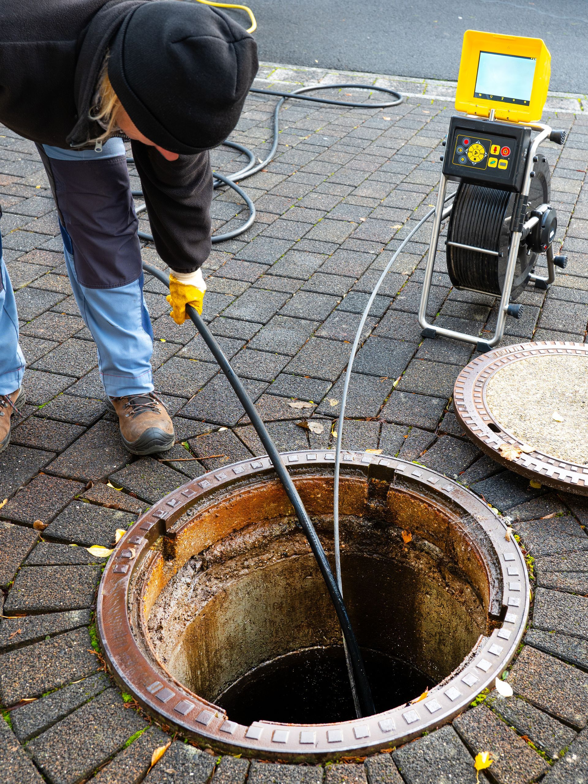 Worker clearing a clogged manhole with a long tool beside a portable pump or vacuum unit.
