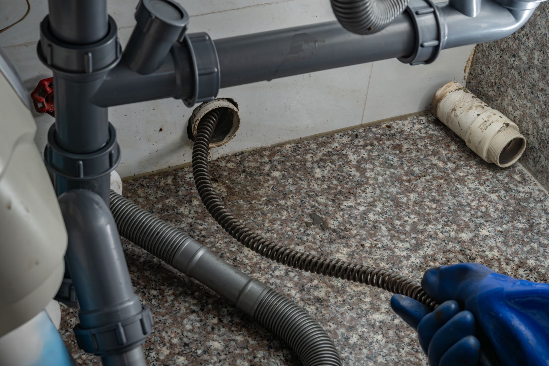 Hand with blue glove holding a flexible drain snake near exposed plumbing under a sink