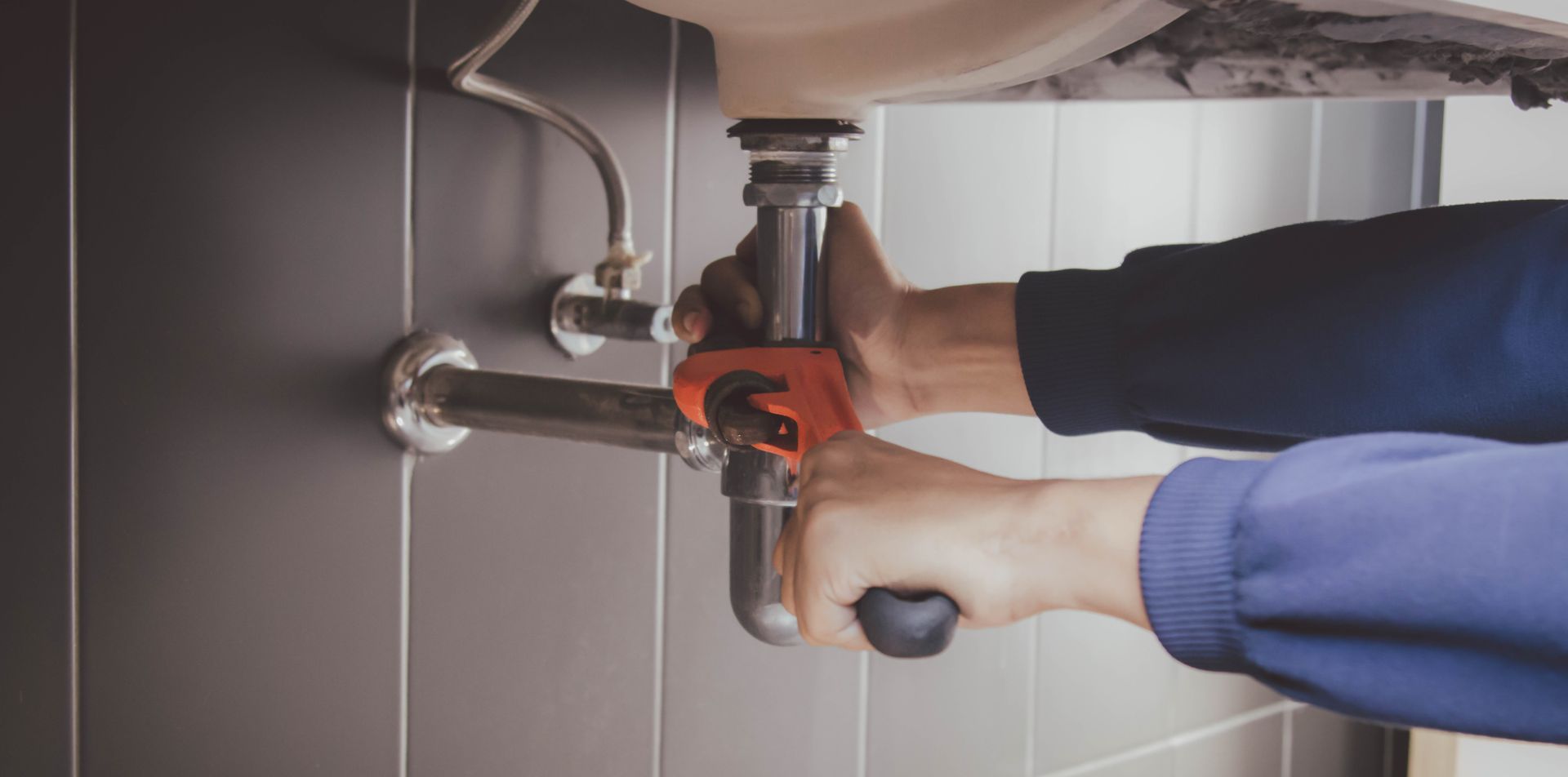 Hands using a wrench to tighten plumbing under a sink, with exposed pipes and a cabinet interior.