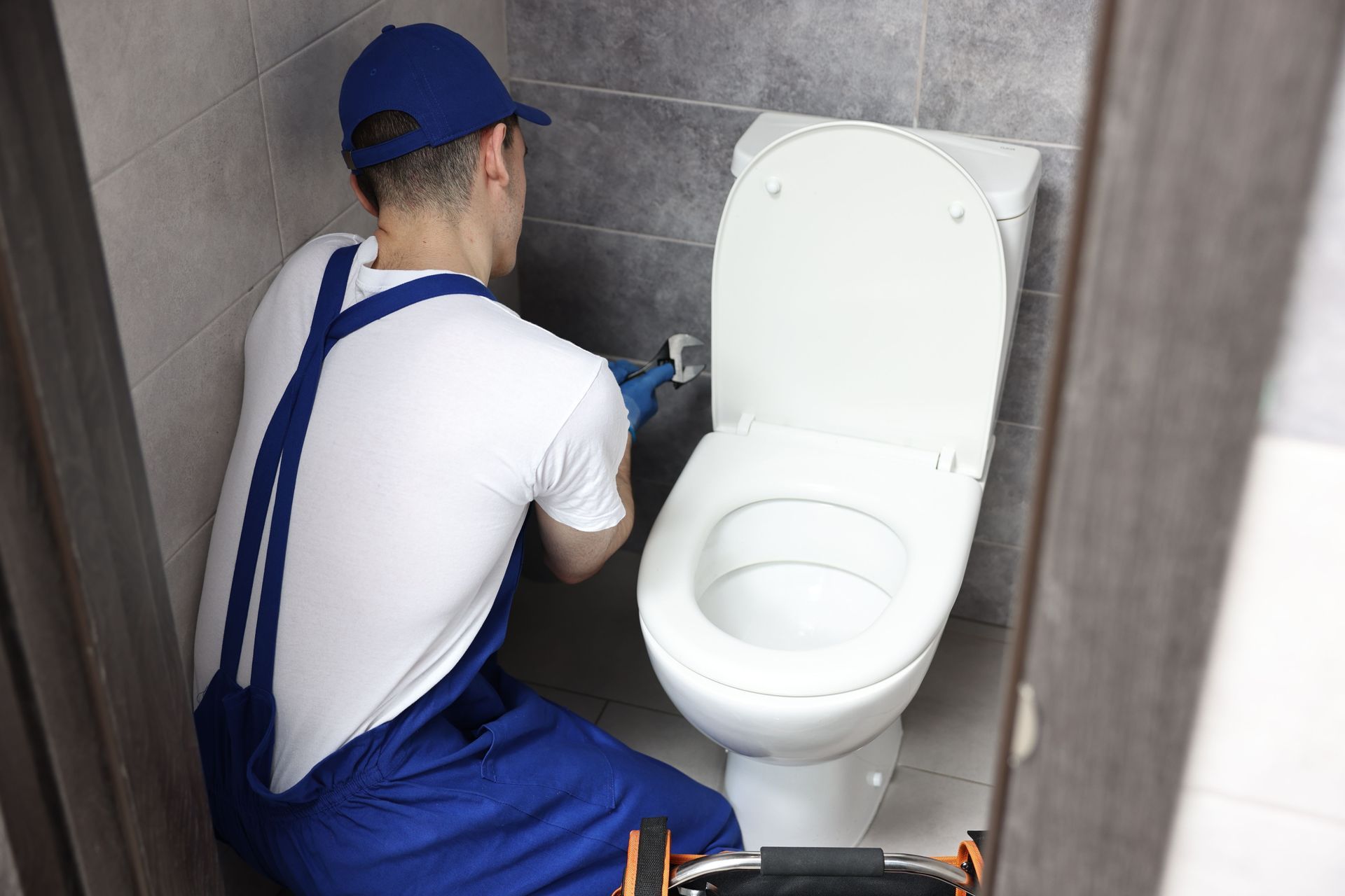 Worker in blue overalls installing a toilet in a tiled bathroom stall