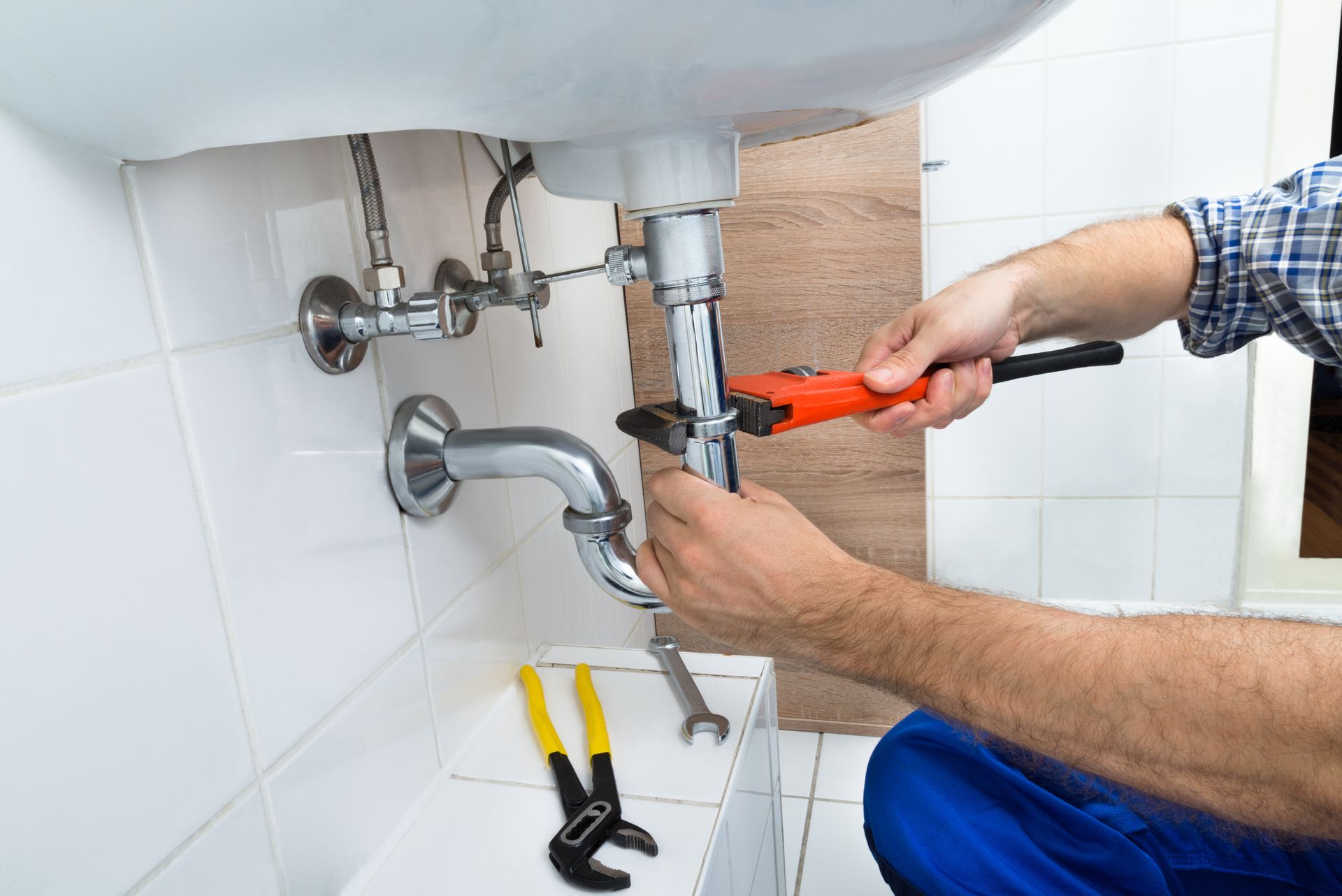 Plumber tightening under-sink pipes with red wrench in a tiled bathroom sink area