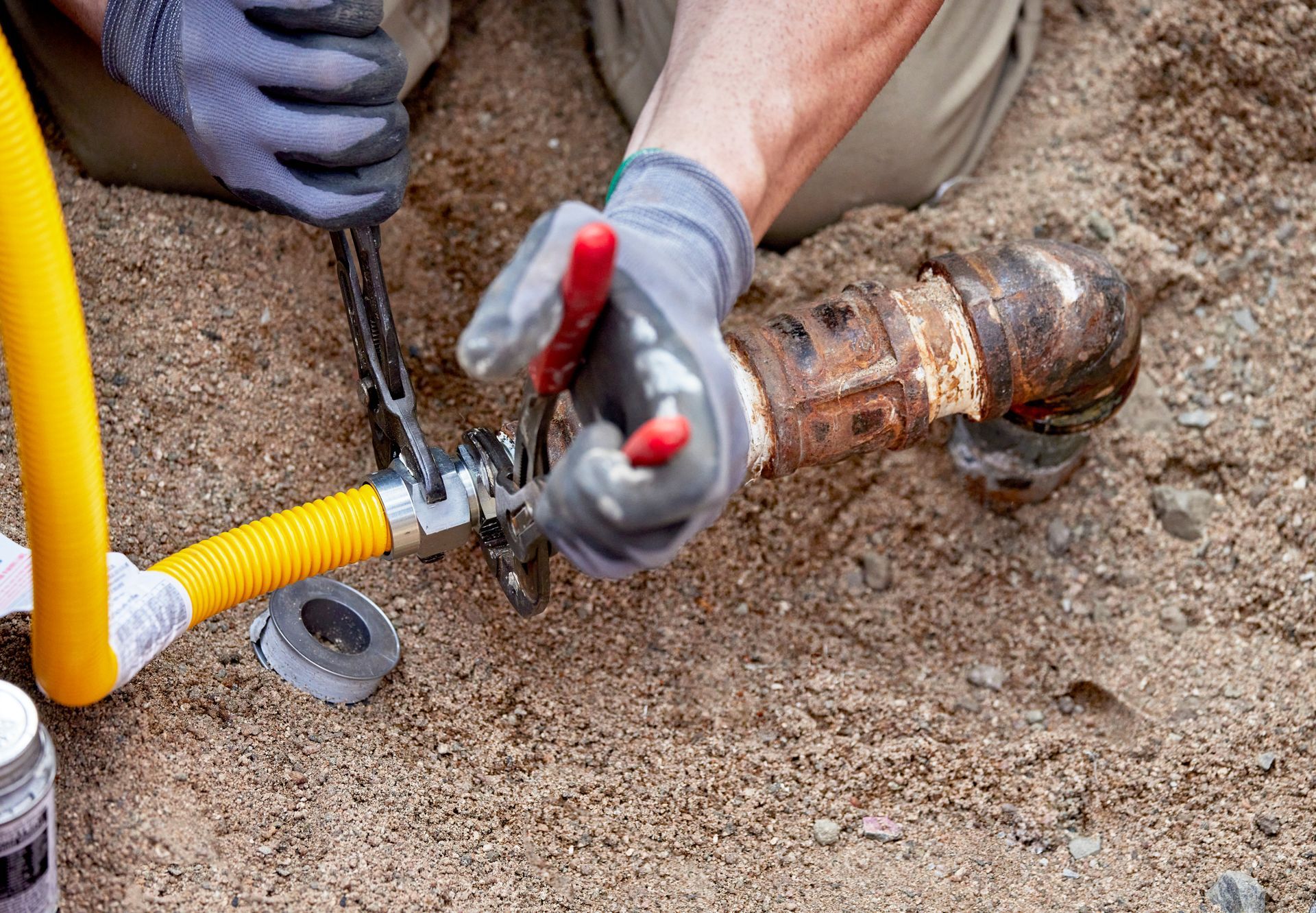 Gloved worker using pliers on a rusty buried pipe next to a yellow hose in dirt