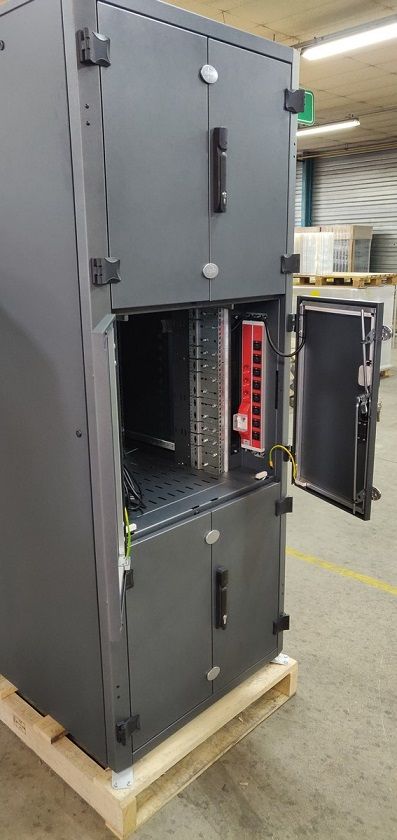 Une armoire électrique gris foncé de style industriel, posée sur une palette en bois dans un entrepôt, sa porte centrale étant ouverte.