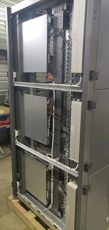 Une baie de serveurs grise avec trois unités métalliques rectangulaires montées et câblage interne, reposant sur une palette en bois.