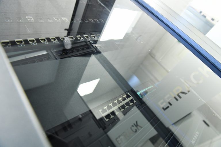 Vue par réflexion à travers une vitrine présentant des équipements serveurs et des panneaux de brassage réseau dans un environnement de bureau.