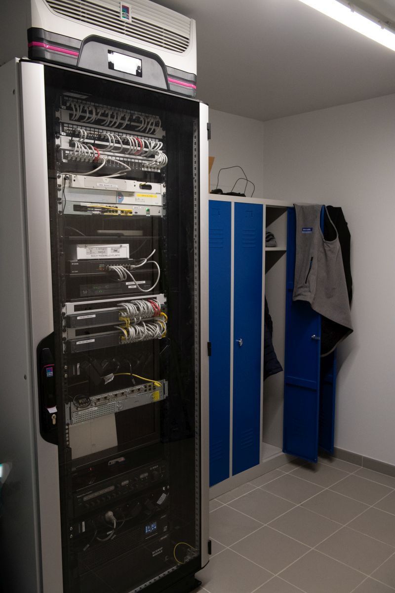 Dans une pièce où se trouvent des casiers de rangement bleus et une veste accrochée sur la droite, se dresse une grande baie serveur à cadre noir.