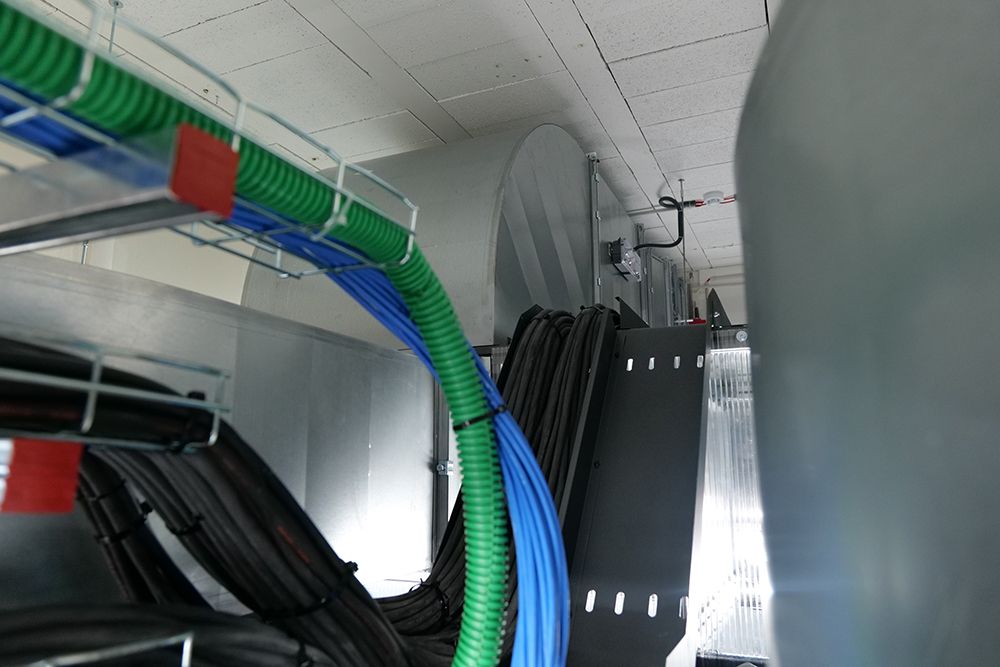 Dans un plafond industriel ou une salle serveur, des conduits verts, des câbles bleus et des fils noirs passent à l'intérieur des chemins de câbles métalliques aériens.