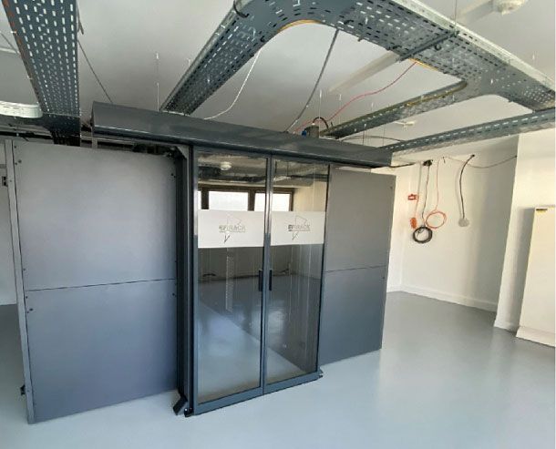 Une baie serveur avec double porte vitrée dans une salle industrielle avec chemins de câbles fixés au plafond et murs blancs.