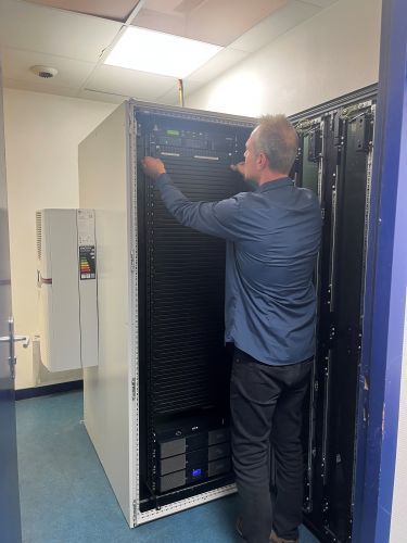 Un technicien travaille sur une baie de serveurs dans un local technique aux murs blancs et au sol bleu.