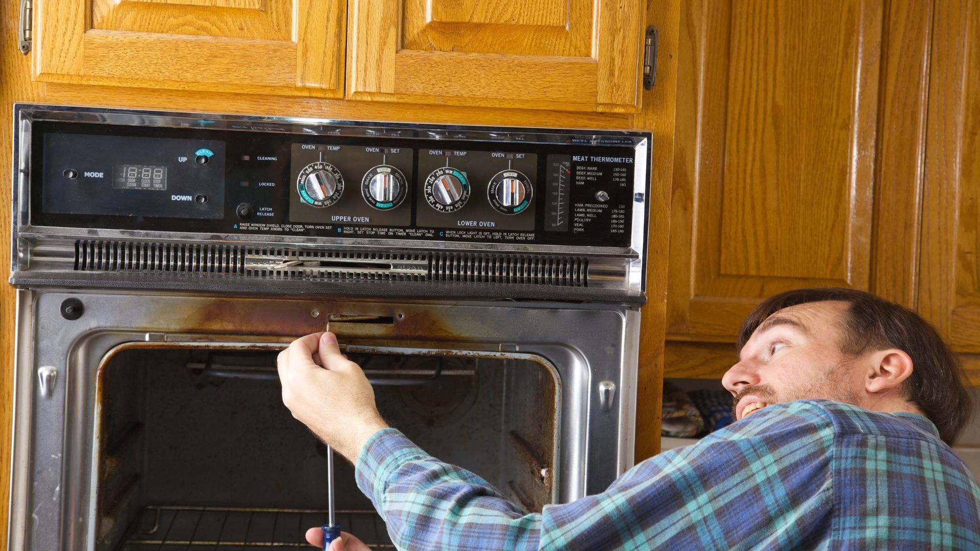 A man is fixing an oven with a screwdriver in a kitchen.
