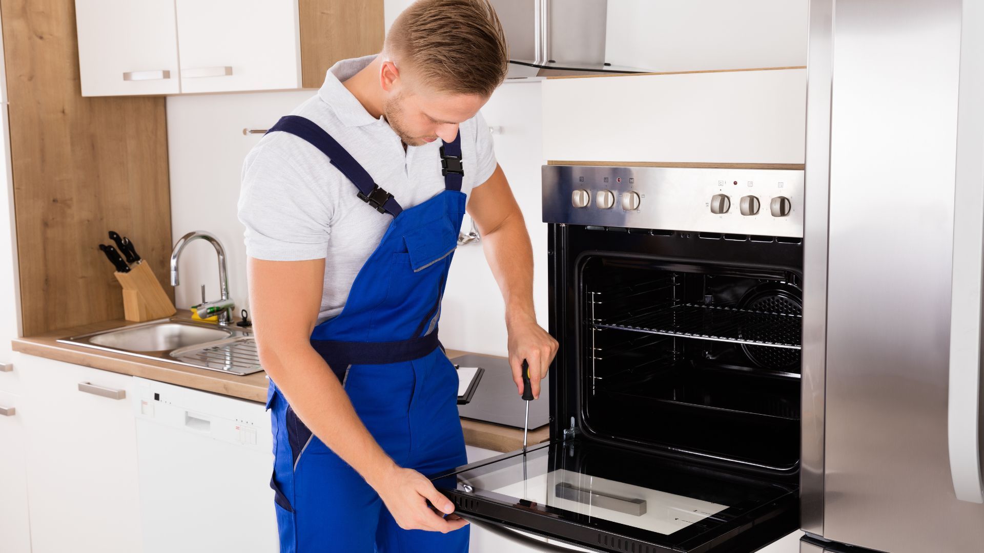 A man in blue overalls is fixing an oven in a kitchen.