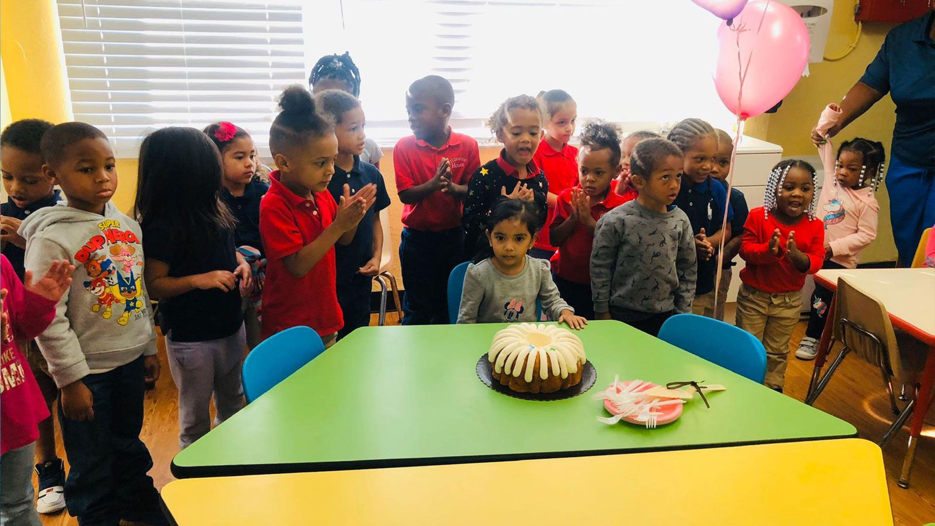A group of children are standing around a table with a cake on it.