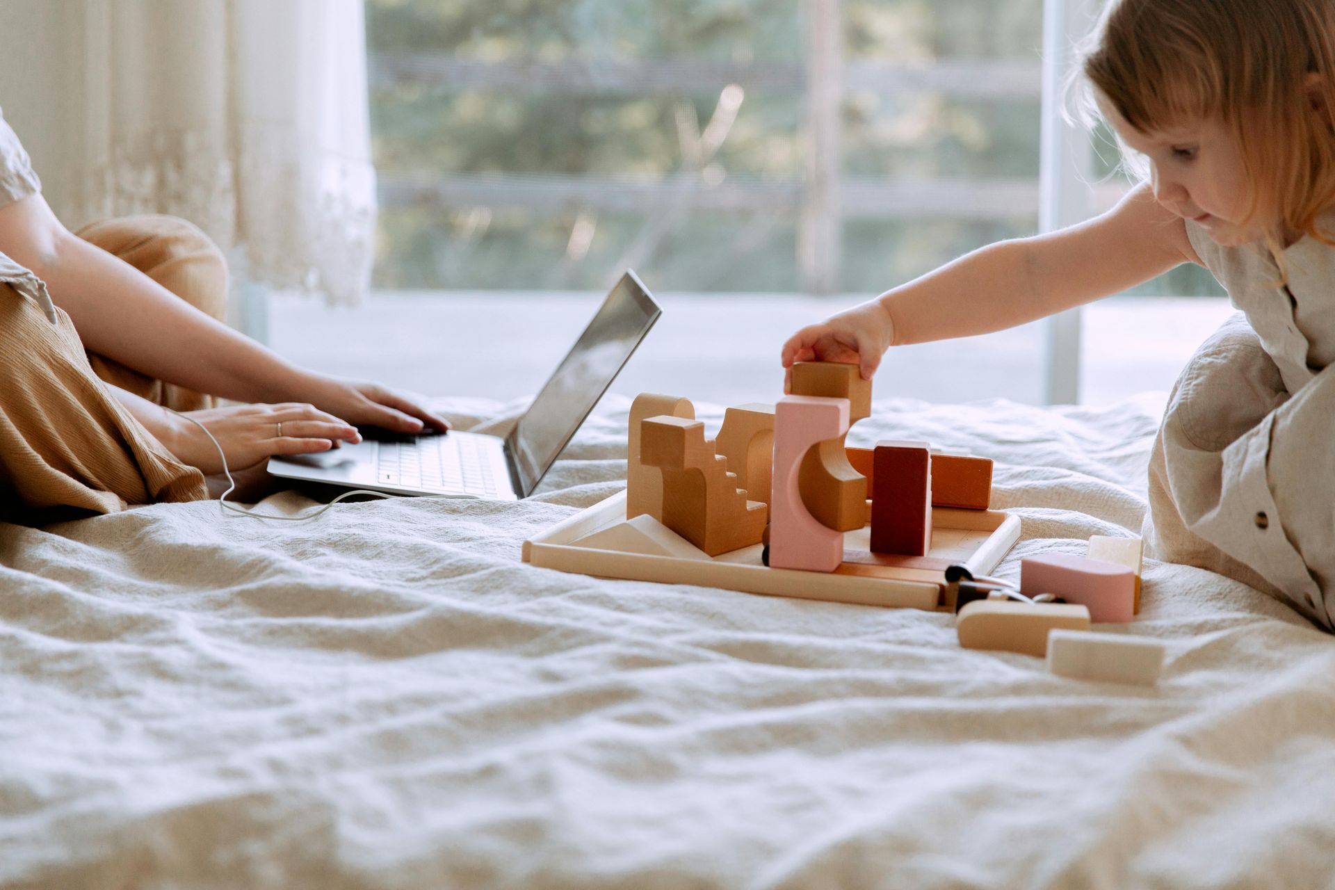 A woman is sitting on a bed with a laptop and a child is playing with wooden blocks.