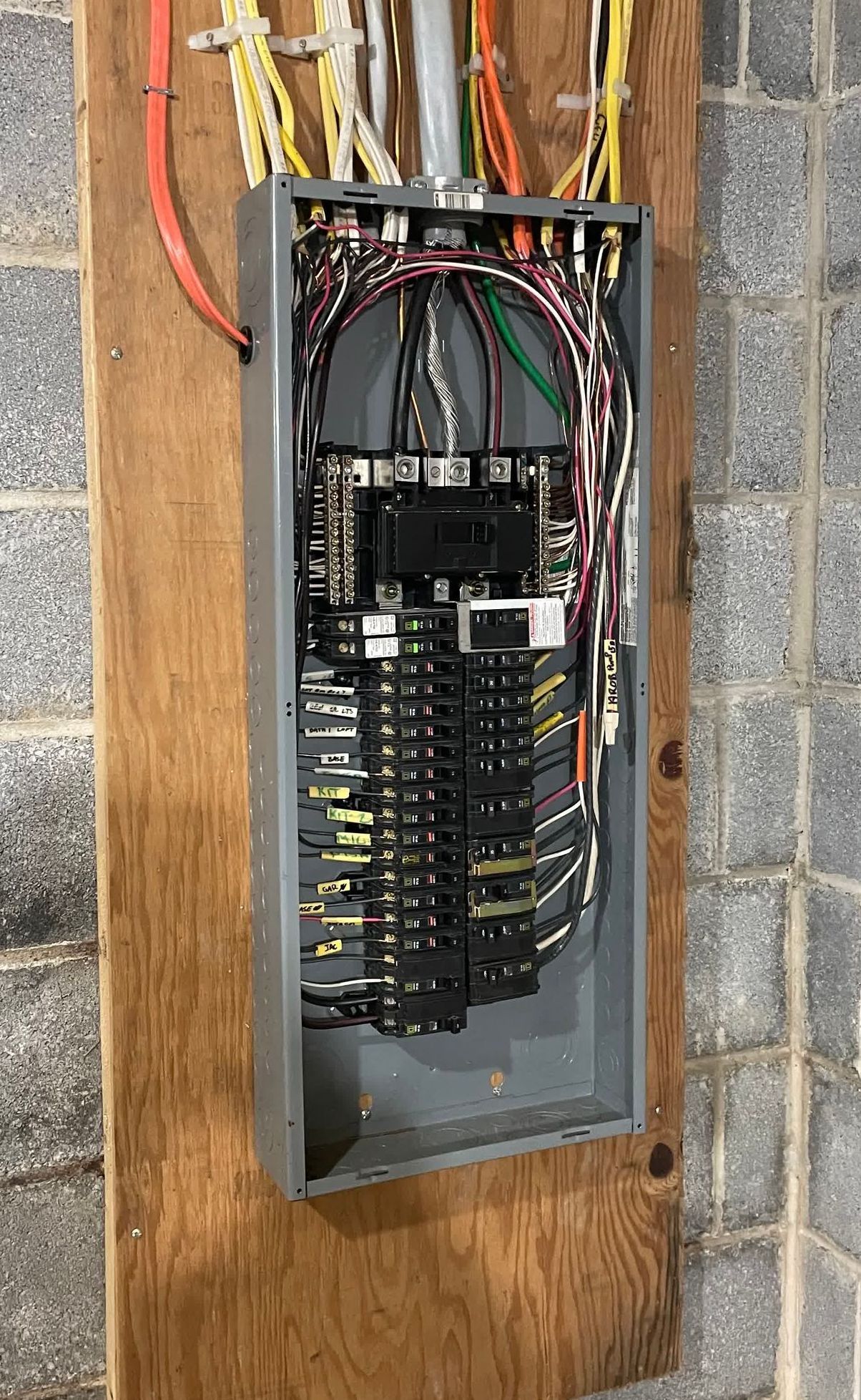 A electrical panel is hanging on a wooden board on a brick wall.