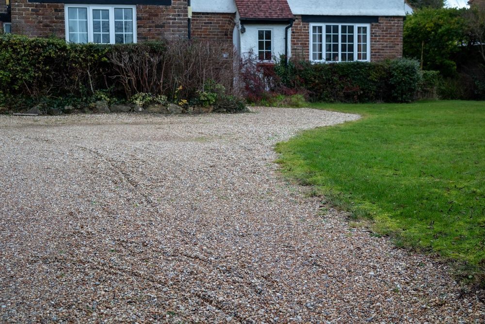 Gravel driveway curving past a green lawn toward a brick house with white windows and hedge borders