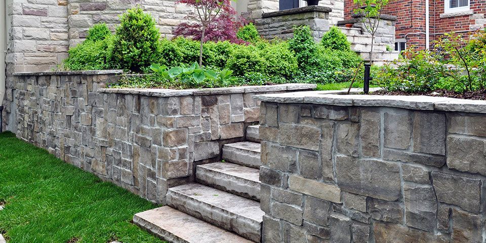 Stone retaining walls with a central set of stone steps leading to a landscaped garden area with shrubs and a lawn.