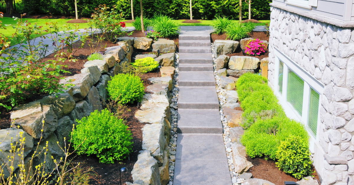 Stone steps flanked by stone retaining walls, green bushes, and the side of a building with windows in a landscaped yard.