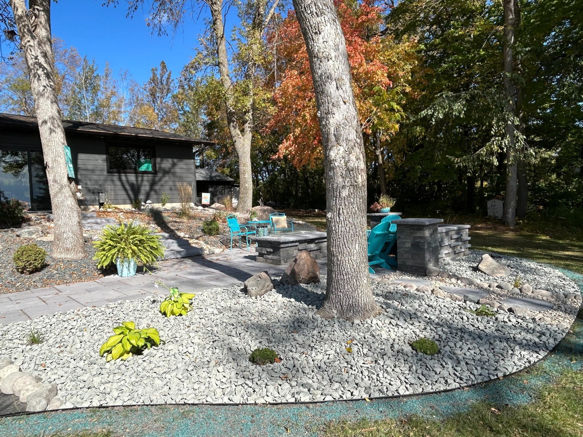 A paved patio with blue chairs and a fire pit sits in front of a dark house, surrounded by light gravel and autumn trees.