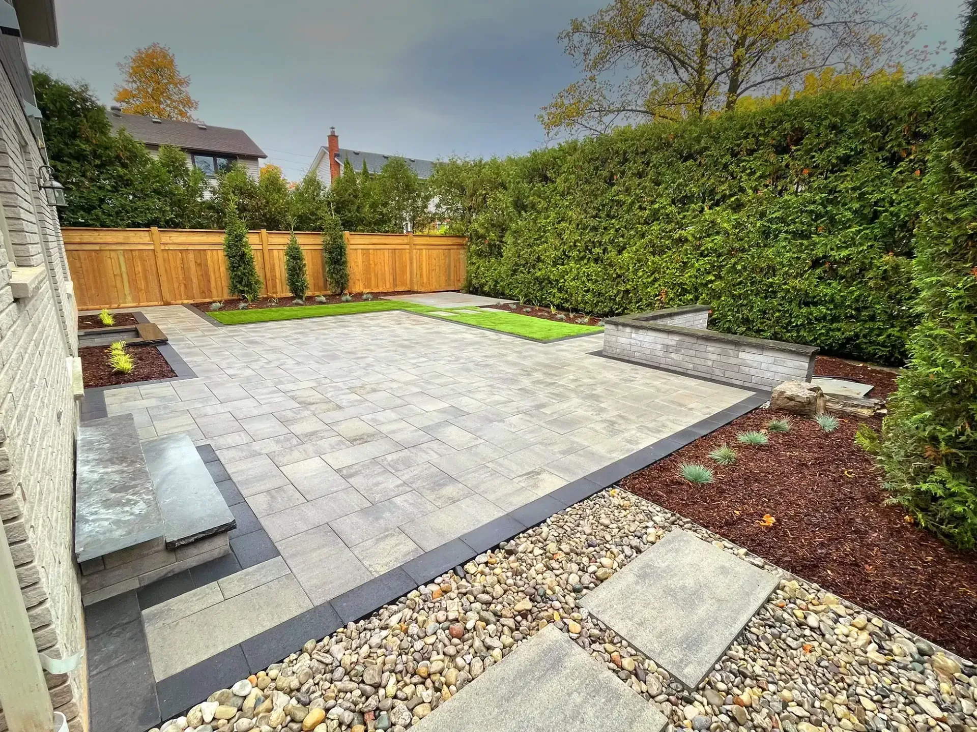 A backyard features a stone paver patio, a raised stone bench, a decorative gravel area, mulch beds, and a wooden fence.