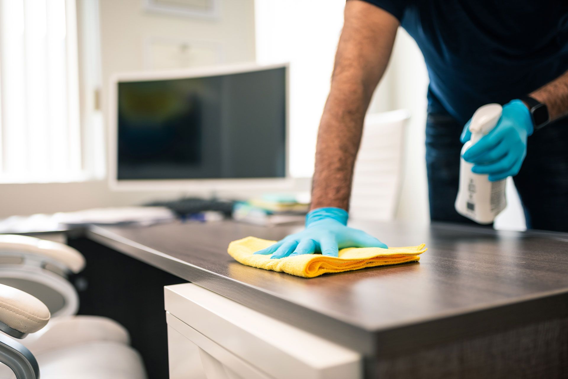 A gloved worker wipes an office desk with a cloth and spray bottle near a computer setup.