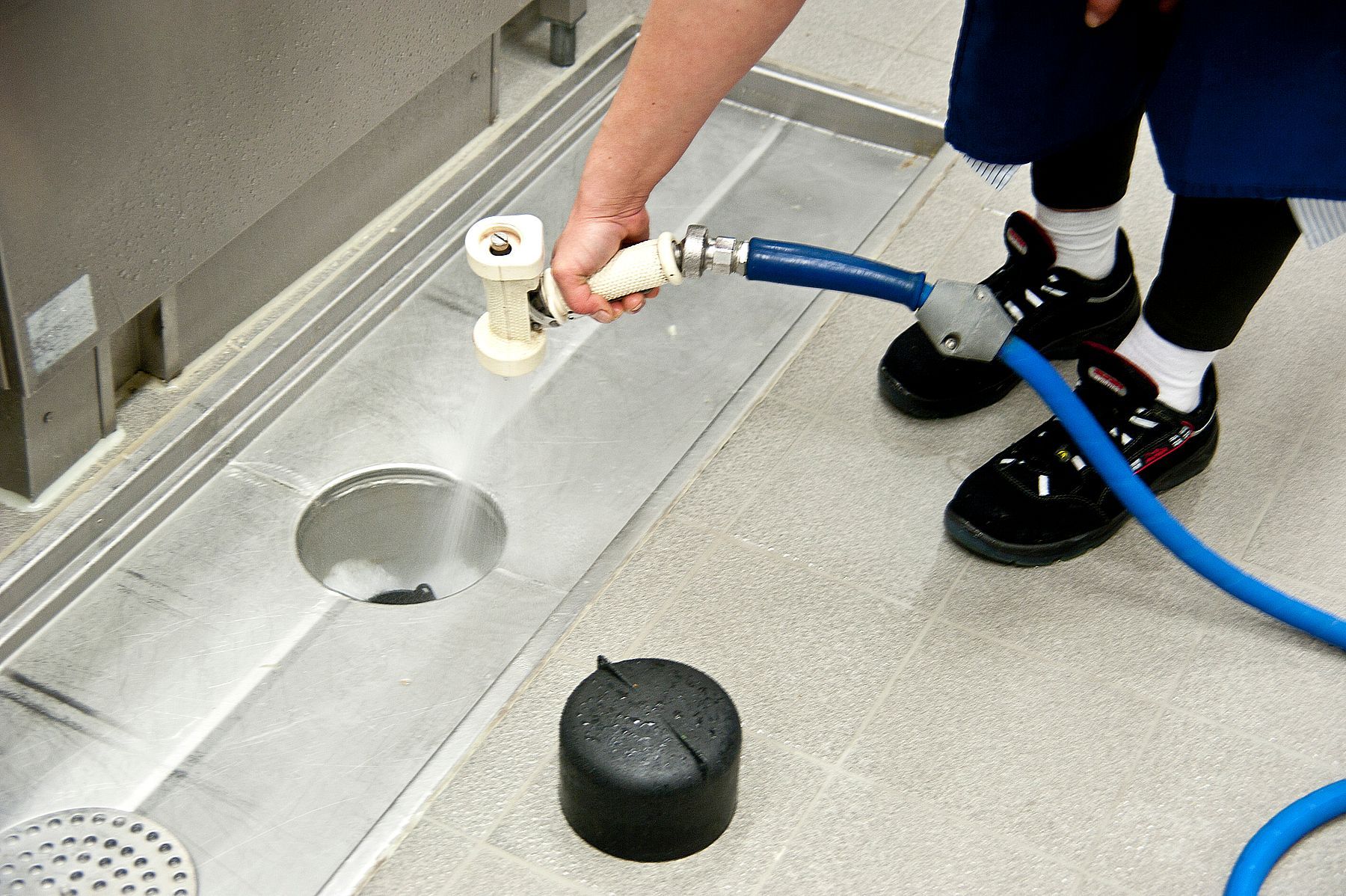 Close-up of a woman’s hand as she uses a pressure nozzle to clean the floor of a commercial kitchen.