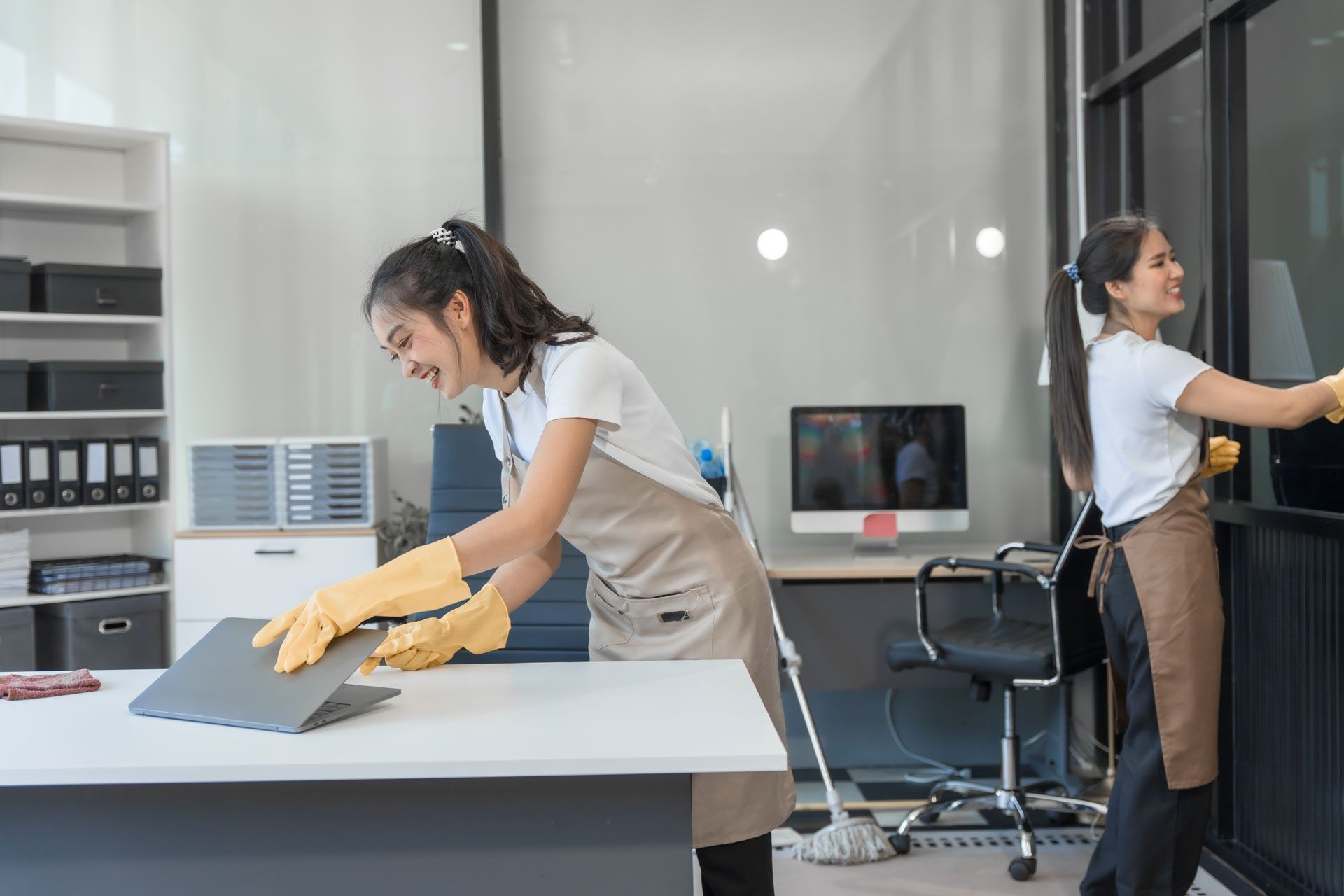 Two cleaners in gloves and aprons clean a desk and glass partition in a modern office. Two cleaners in gloves and aprons clean a desk and glass partition in a modern office.