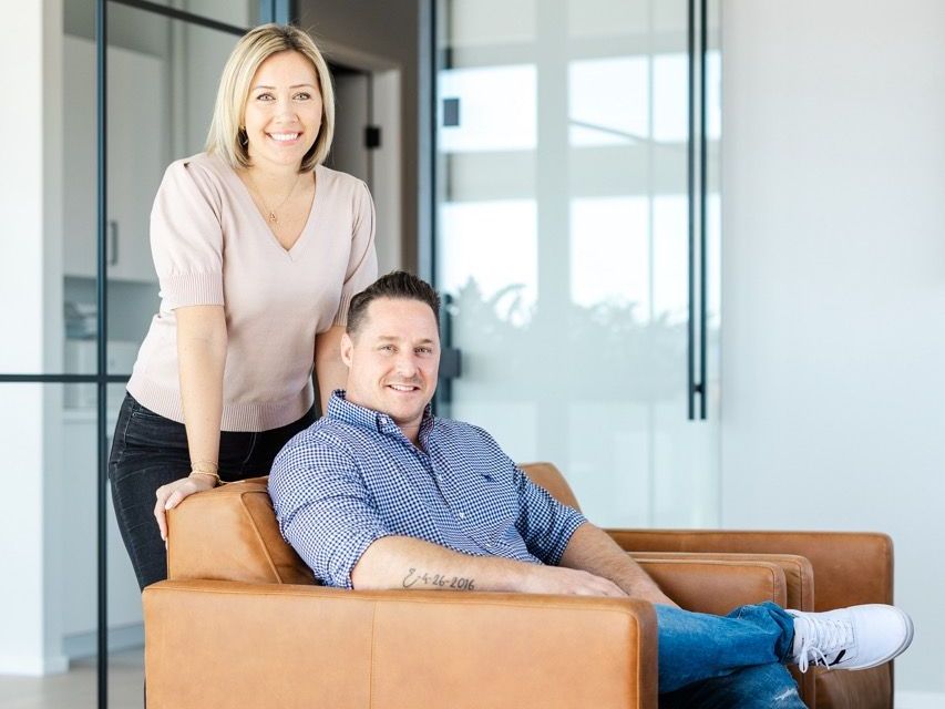 Woman leaning on man seated in brown leather chair, smiling. Glass office setting.