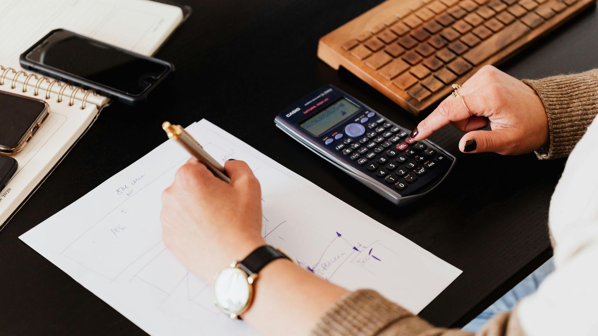 Person using a calculator and writing on paper at a desk, near a phone and keyboard.